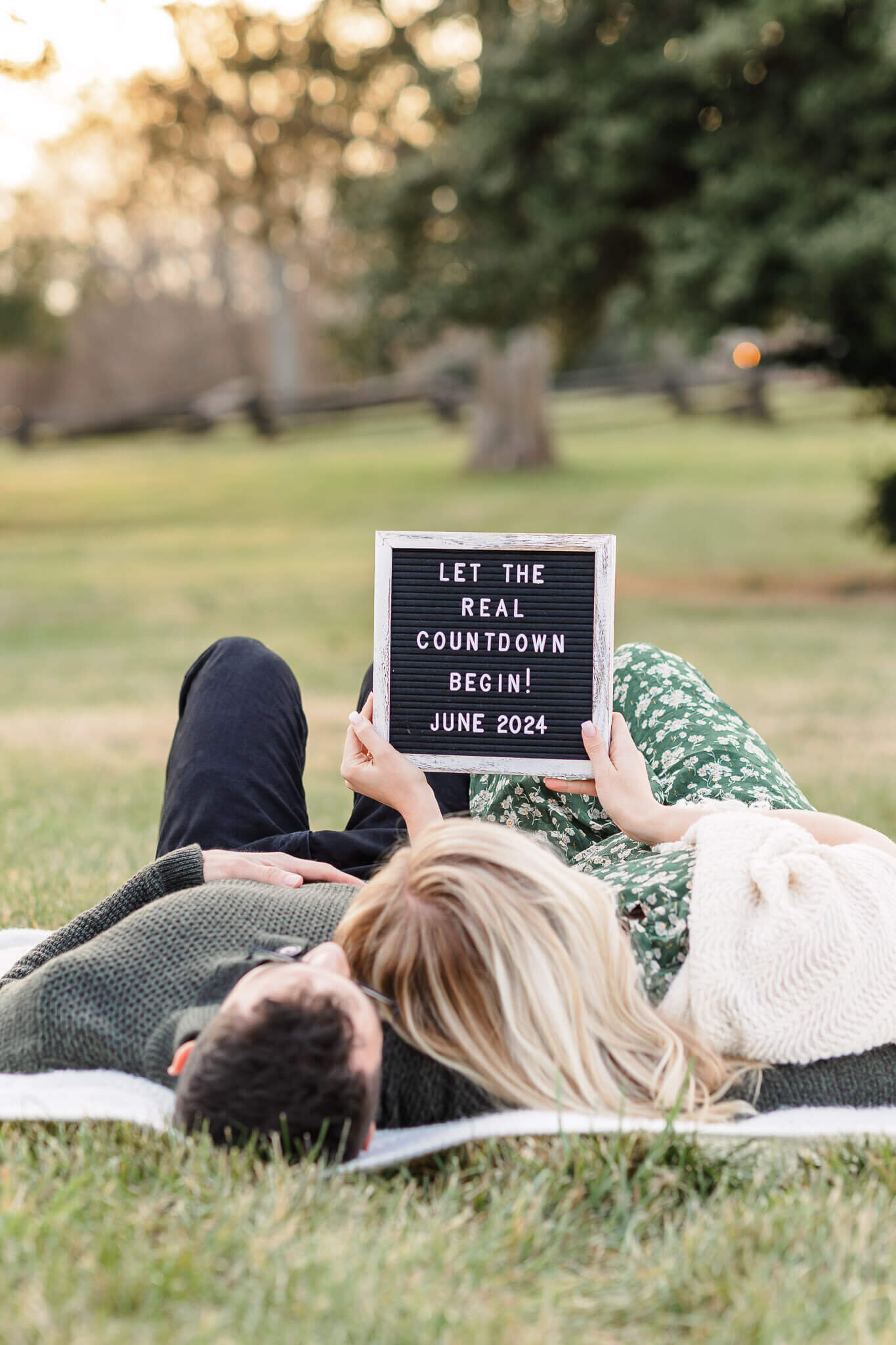 A mom and dad laying in the grass and holding up a sign announcing their pregnancy.