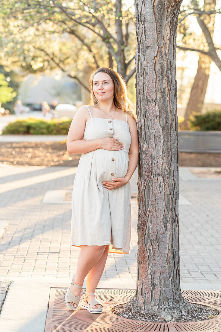 A pregnant woman wearing a cream colored sundress holds her belly as she leans against a tree at BYU, Provo at sunset