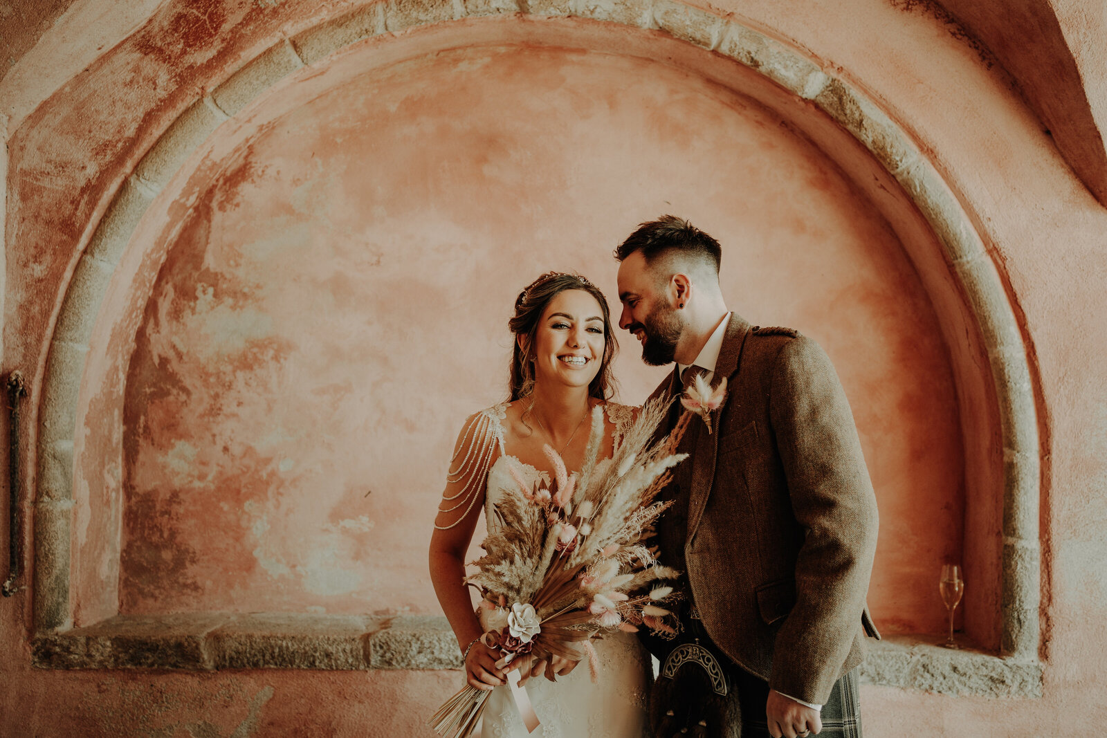 bride in white dress holding bridal bouquet and groom wearing kilt and tweed jacket in front of pink stone arch natural aberdeen wedding photography