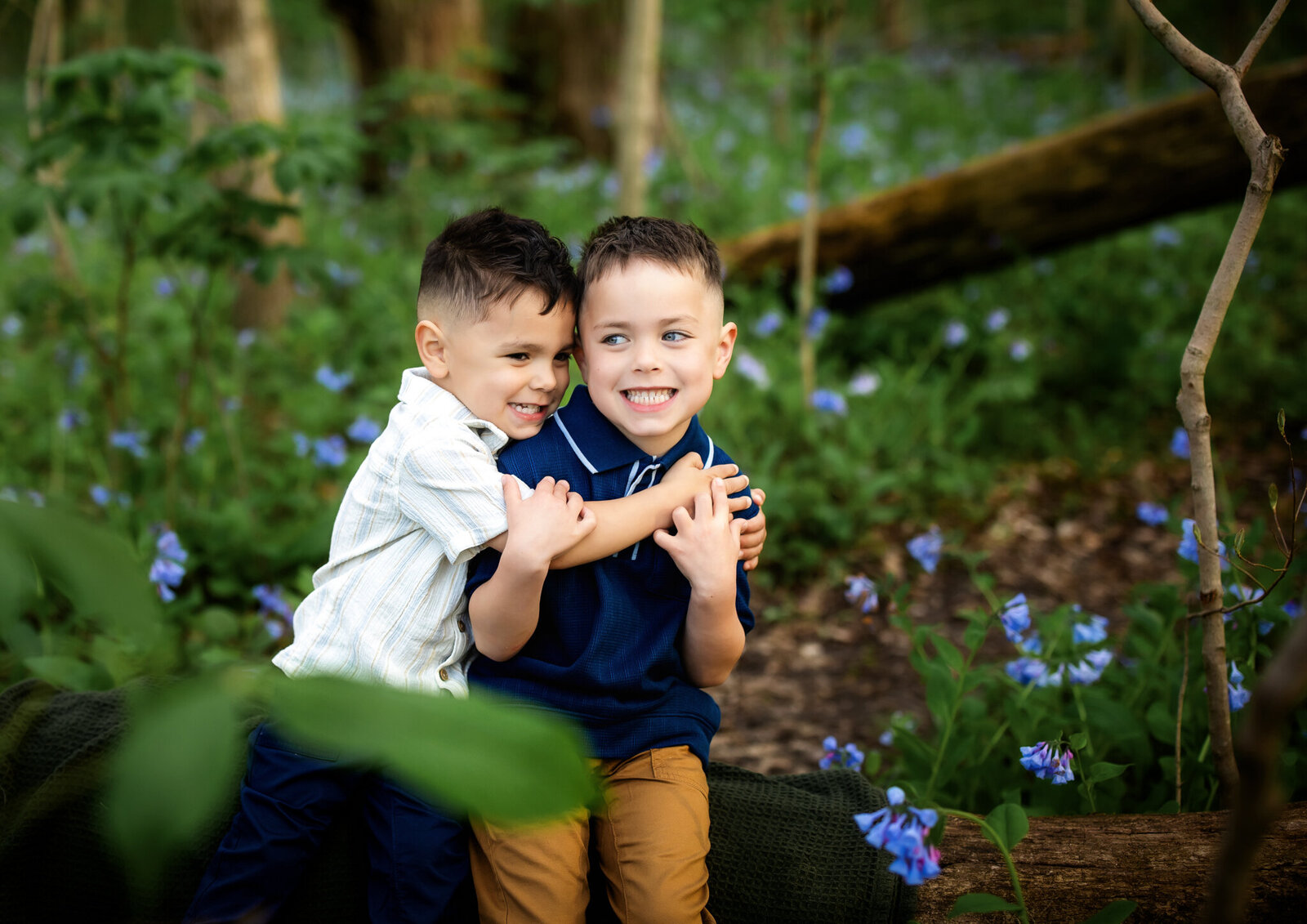 Two young boys sit close together on a log in a lush, green forest filled with blue wildflowers. Smiling and hugging, they look happy and playful—an ideal moment for outdoor portraits by a Bloomington family photographer.