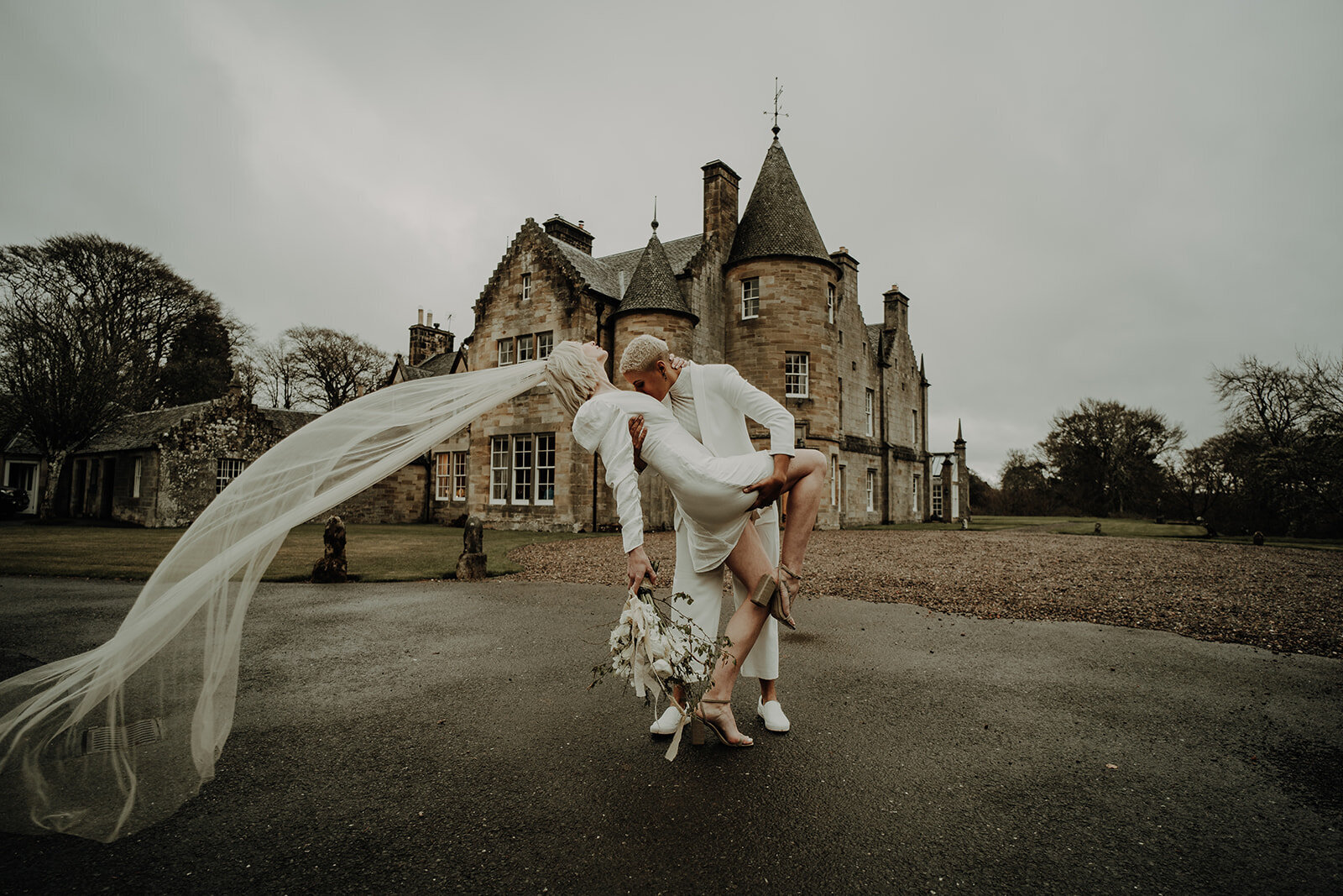 two brides in white outfits  one leaning back as her veil is swept in the wind creative alternative wedding photographer scotland