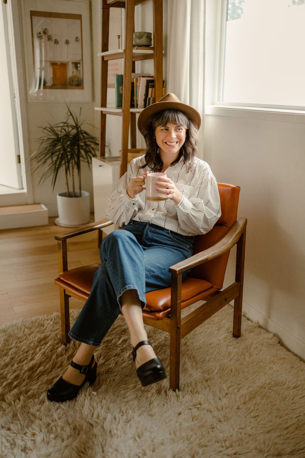 Woman sitting in chair wearing a hat and holding a coffee cup.