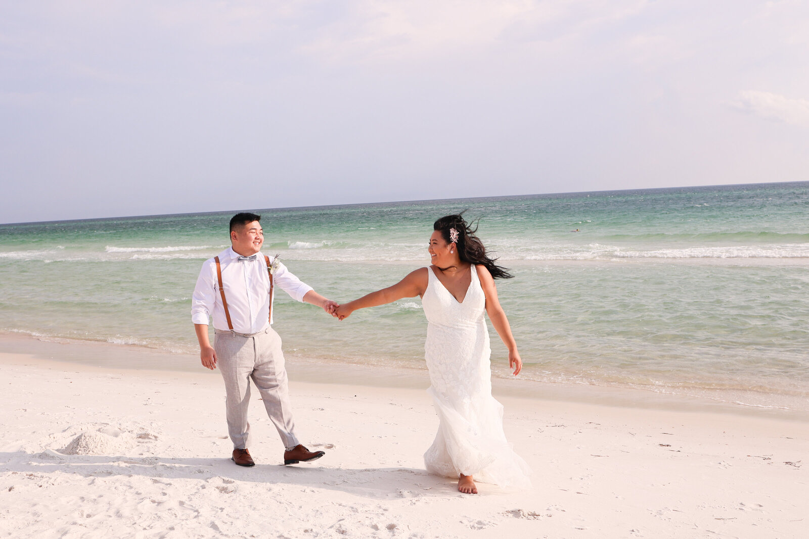 bride and groom holding hands while walking on the beach. Bride is wearing a v neck lace wedding dress and groom is wearing gray pants, while button down shirt and brown suspenders. 