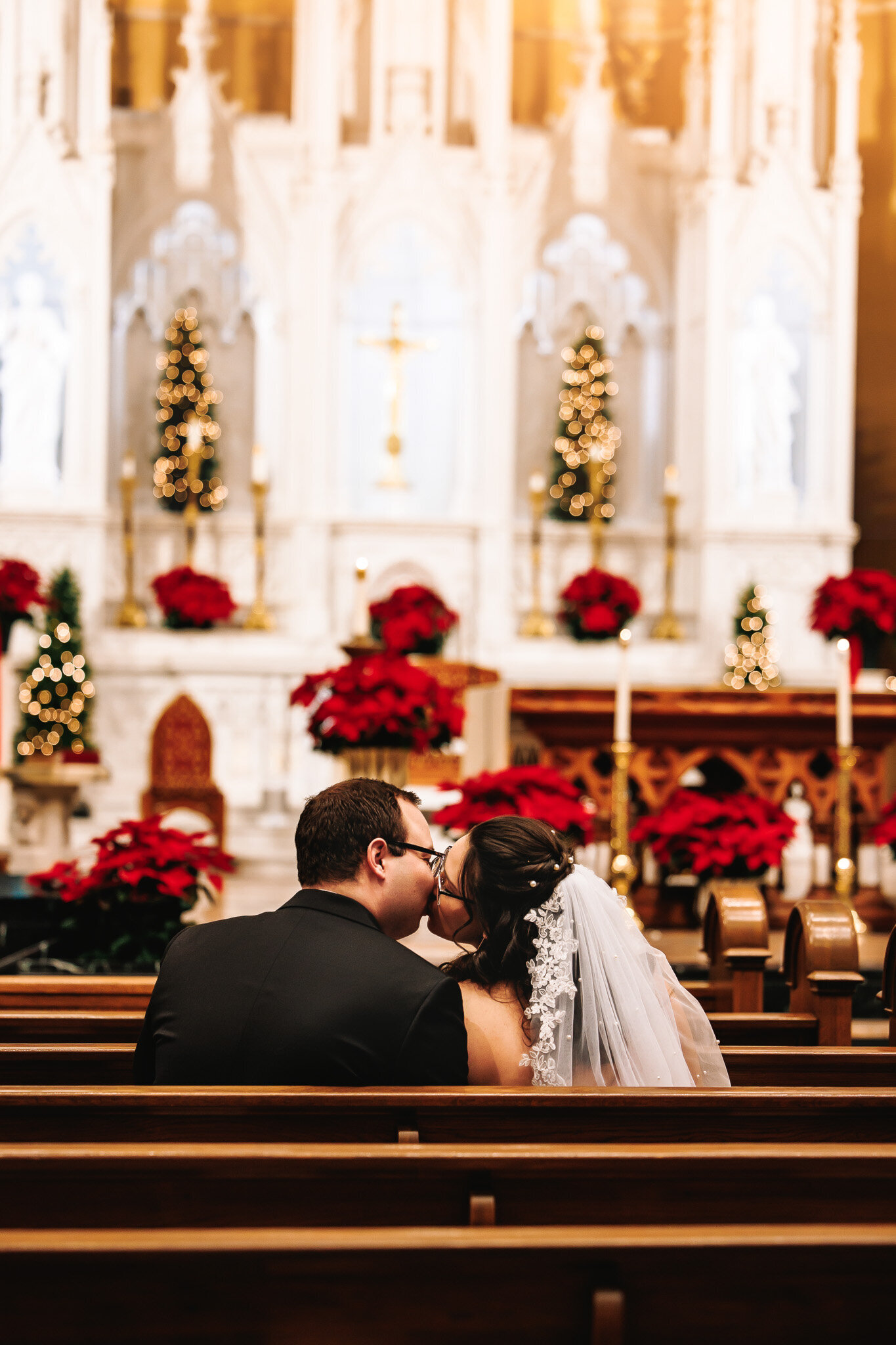 A bride and groom share a kiss sitting in the pews of the church. An altar surrounded by poinsettias and Christmas lights in the background 
