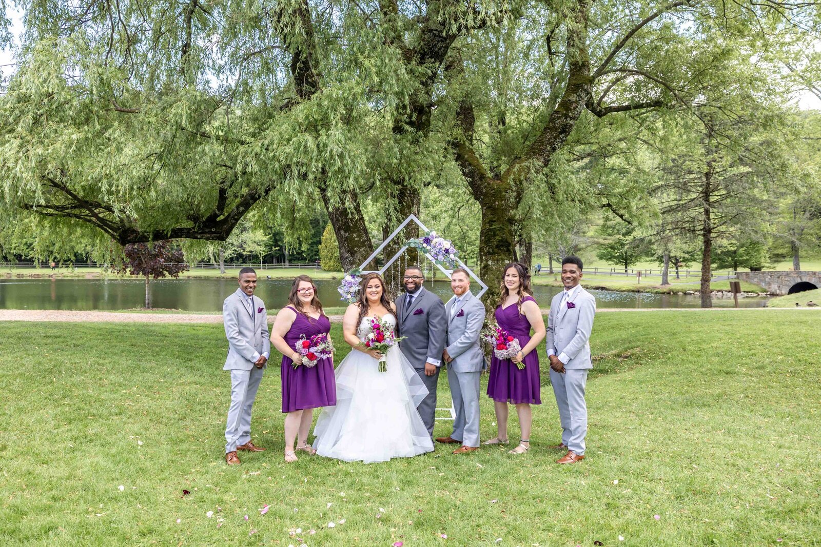 The bride and groom pose with their wedding party by a lakeside arbor adorned with purple florals, captured during an outdoor Chetola Resort wedding, in beautiful Blowing Rock, NC blending vibrant colors and rustic charm.