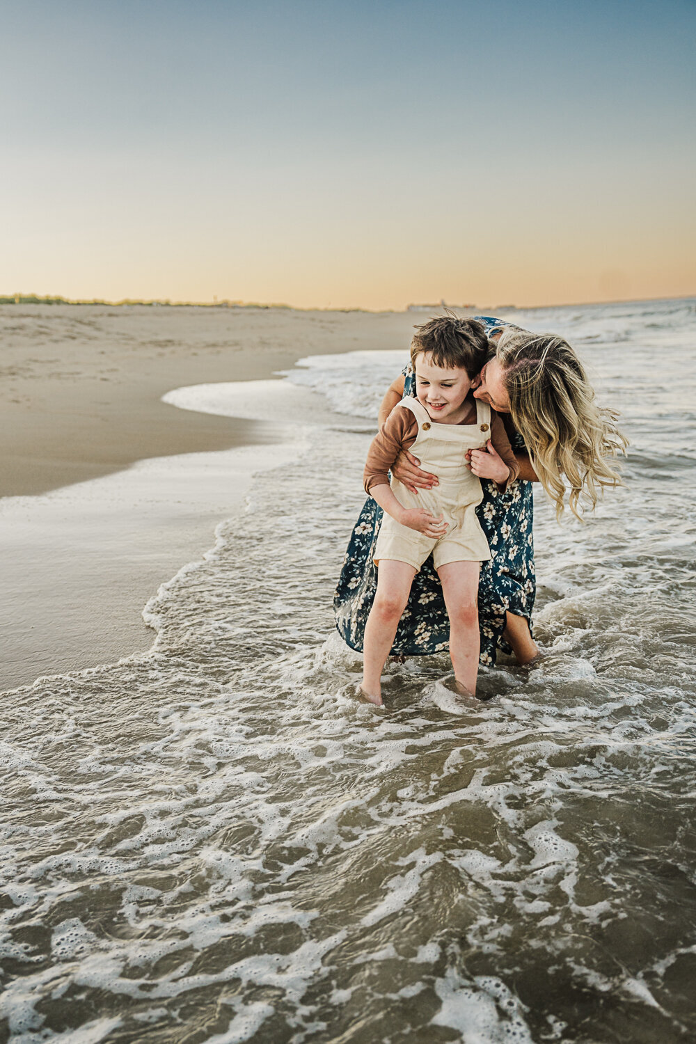 mom snuggles toddler boy in overalls in the waves of the beach