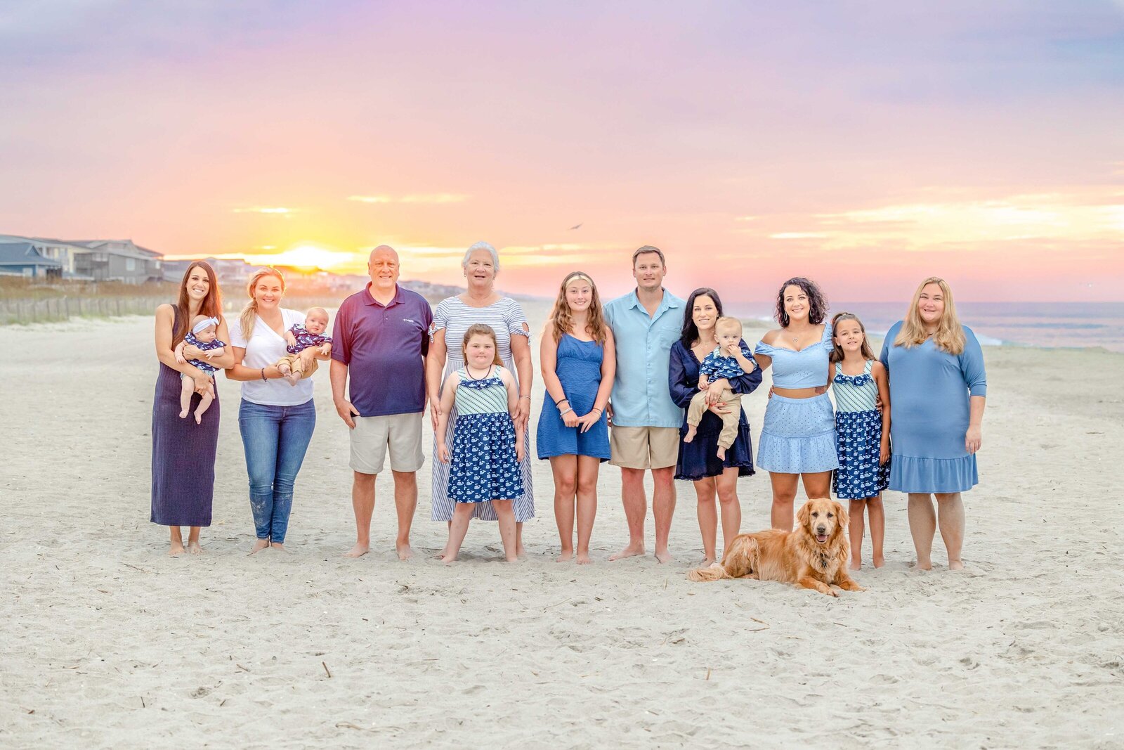 A large family, including a golden retriever, poses on Ocean Isle Beach at sunset. Dressed in coordinated blue and white outfits, they capture the warmth of a family vacation. Perfect for North Carolina family photography, highlighting vibrant sunset gatherings.