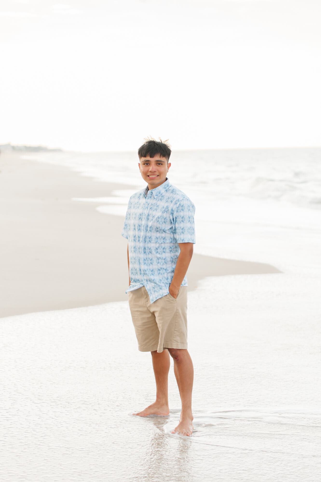 Senior boy wearing blue stands near the ocean for senior portraits