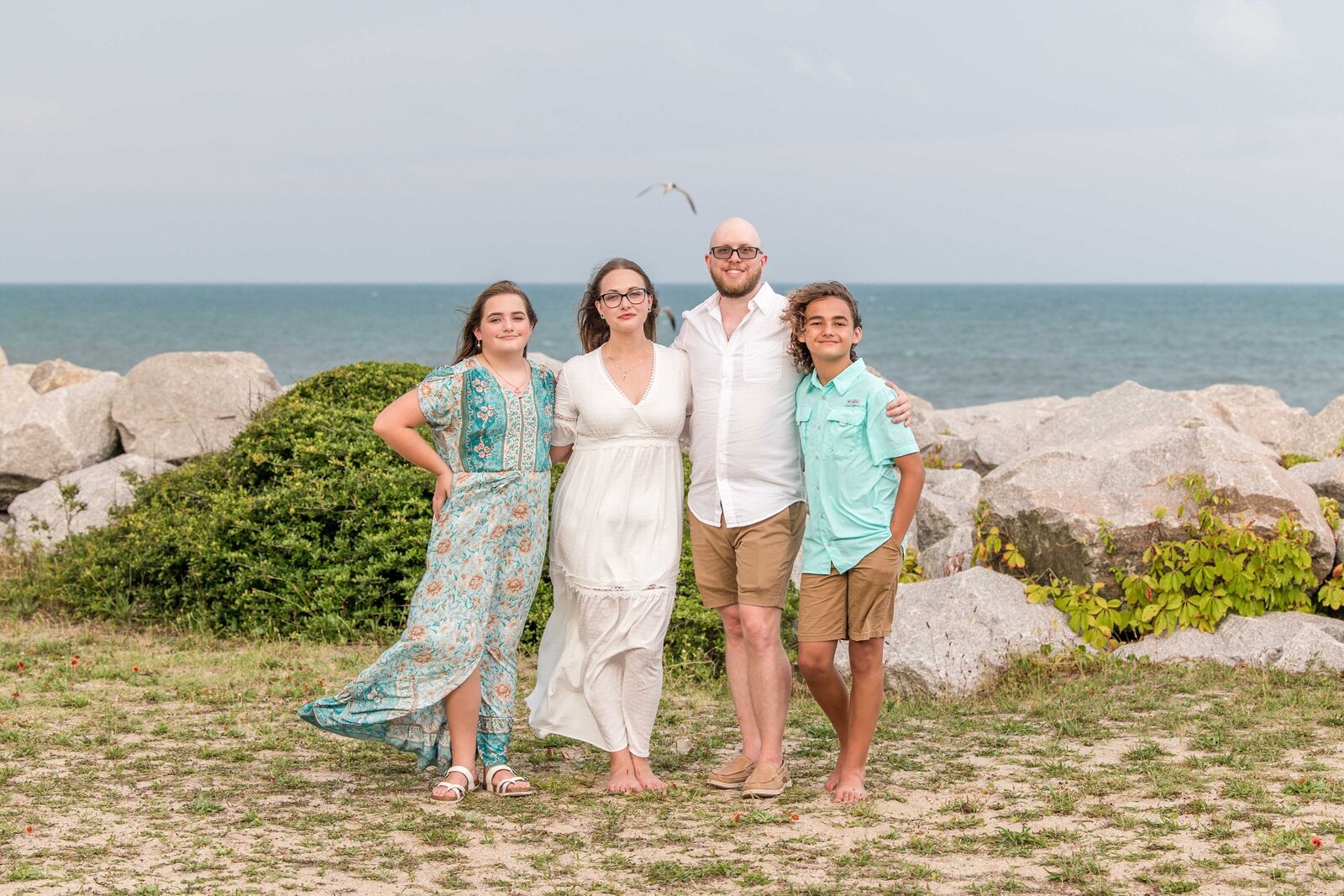 A family of four poses near the rocky coastline, with the ocean as a backdrop. Dressed in casual summer attire, their smiles reflect a relaxed and joyful atmosphere. Perfect for professional family photography in NC, highlighting natural coastal beauty.