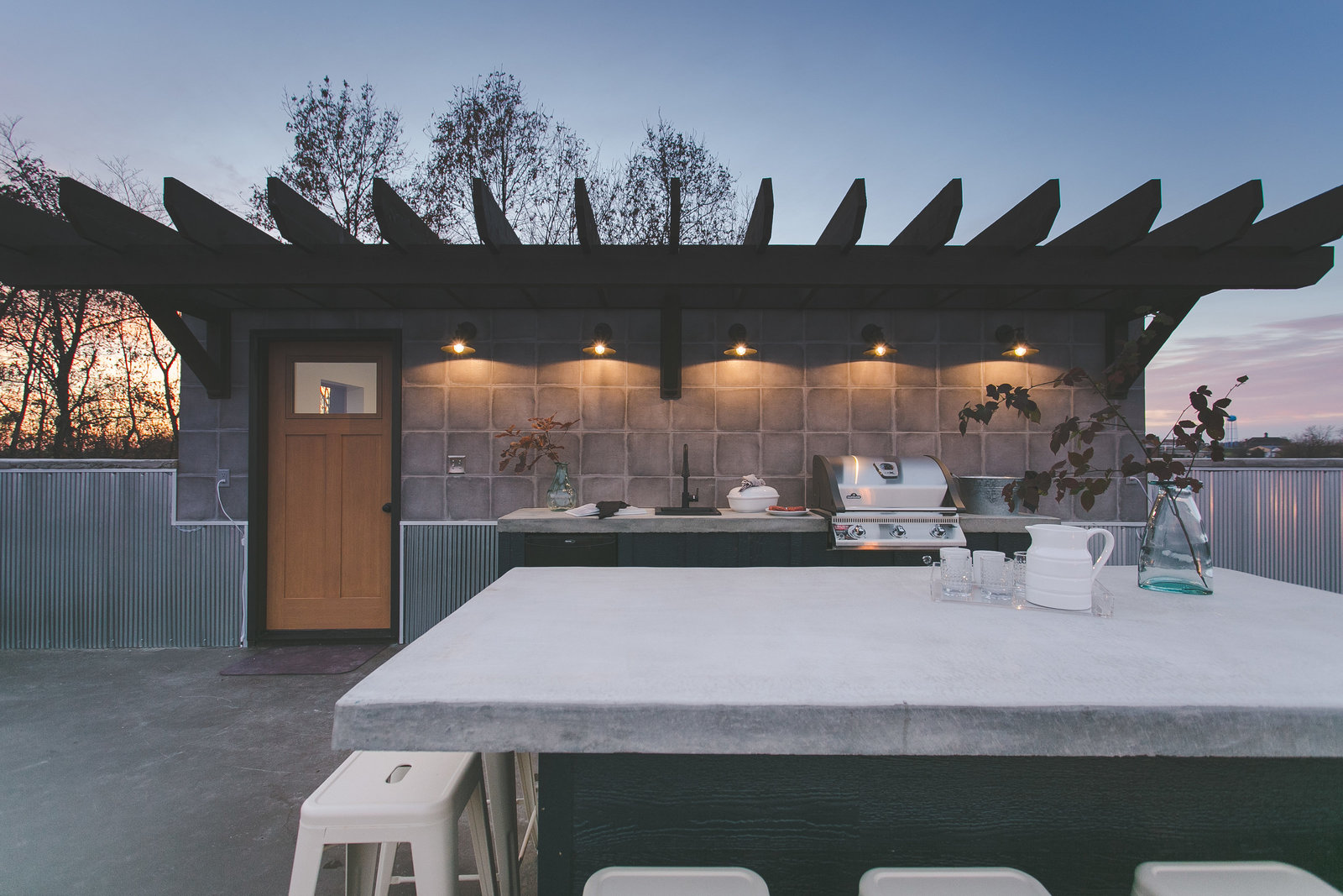 Modern BnB rooftop kitchen design with concrete countertops, black exterior sconces and white barstools. Custom mid tone wood door softens the look of the industrial metal siding surrounding the rooftop.