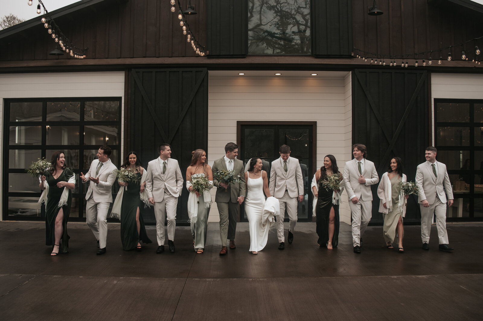 Wedding party walking together at a rustic venue, featuring groomsmen in light gray suits and bridesmaids in deep green dresses, posed against large dark wooden barn doors for a charming outdoor wedding photoshoot at Sage and Social in Mulino, Oregon.