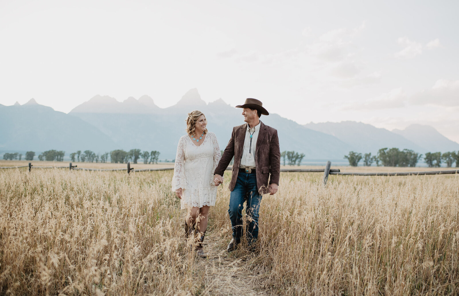 wyoming elopement with cowboy groom holding hands with his bride as they walk through a dried grass field together hodling hands with the grand tetons in the distance as the sun sets through the peeks