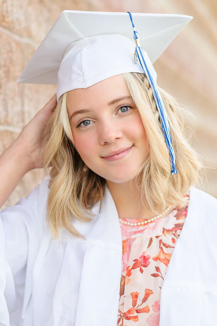 A closeup of a high school senior girl wearing a white graduation cap and gown standing against a brick wall at North Lake Park in Lehi