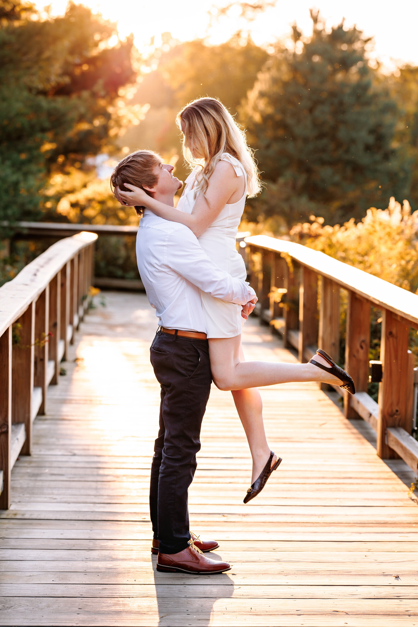 Man lifts up his fiancé along a wooden walkway with the golden glow of the sun in the background