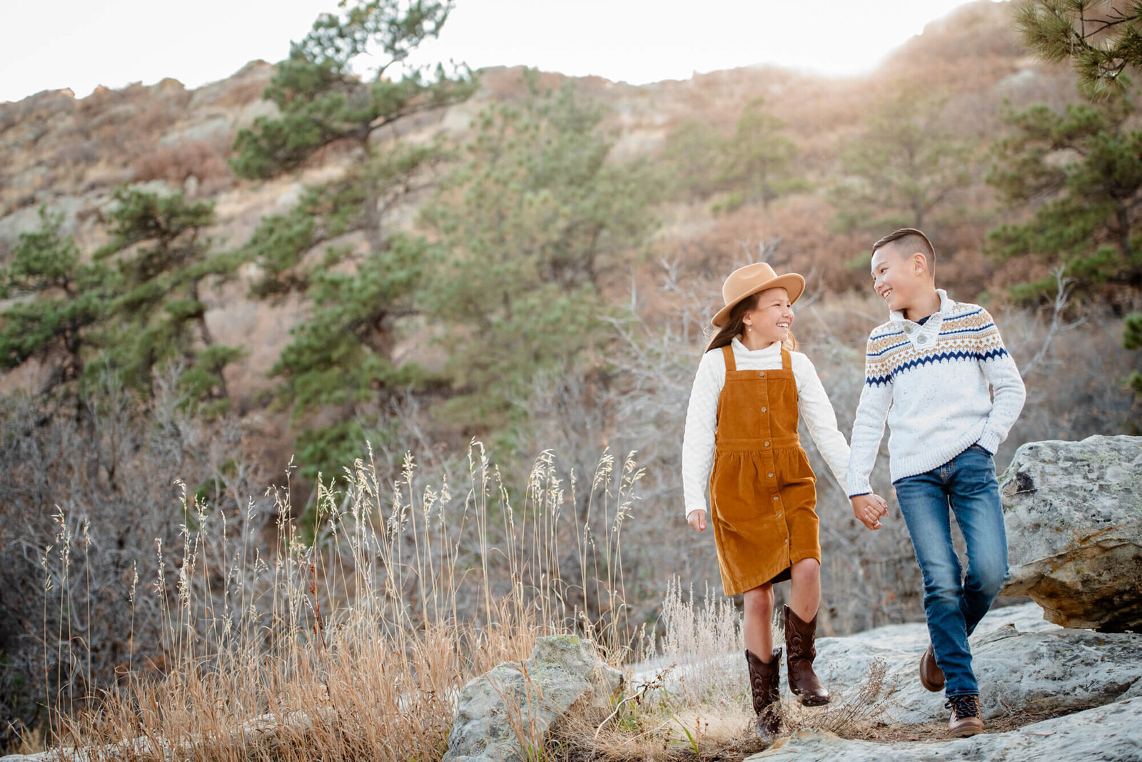 A young brother and sister walk a rocky mountain trail holding hands at sunset for a Denver family photographer