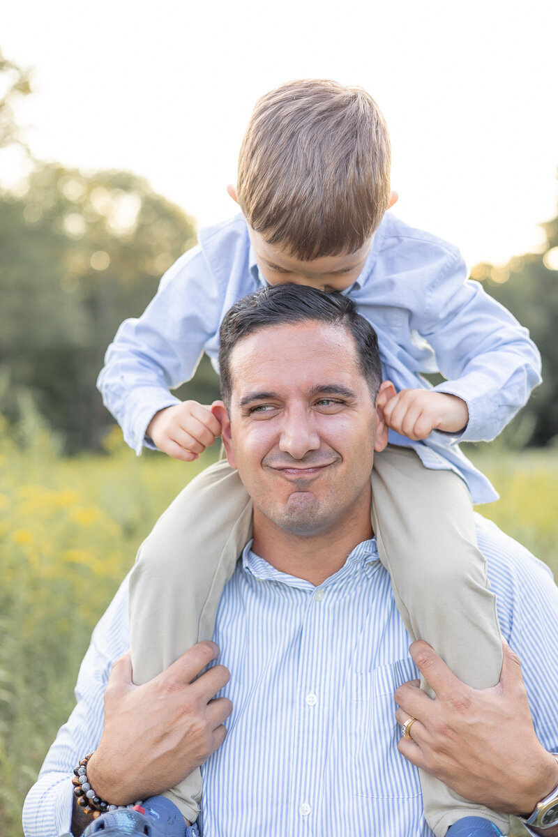 Candid photo of a dad making a silly face while his young son kisses his head from atop his shoulders—photographed by Westchester family photographer Nicole during a golden hour session in a wildflower meadow.
