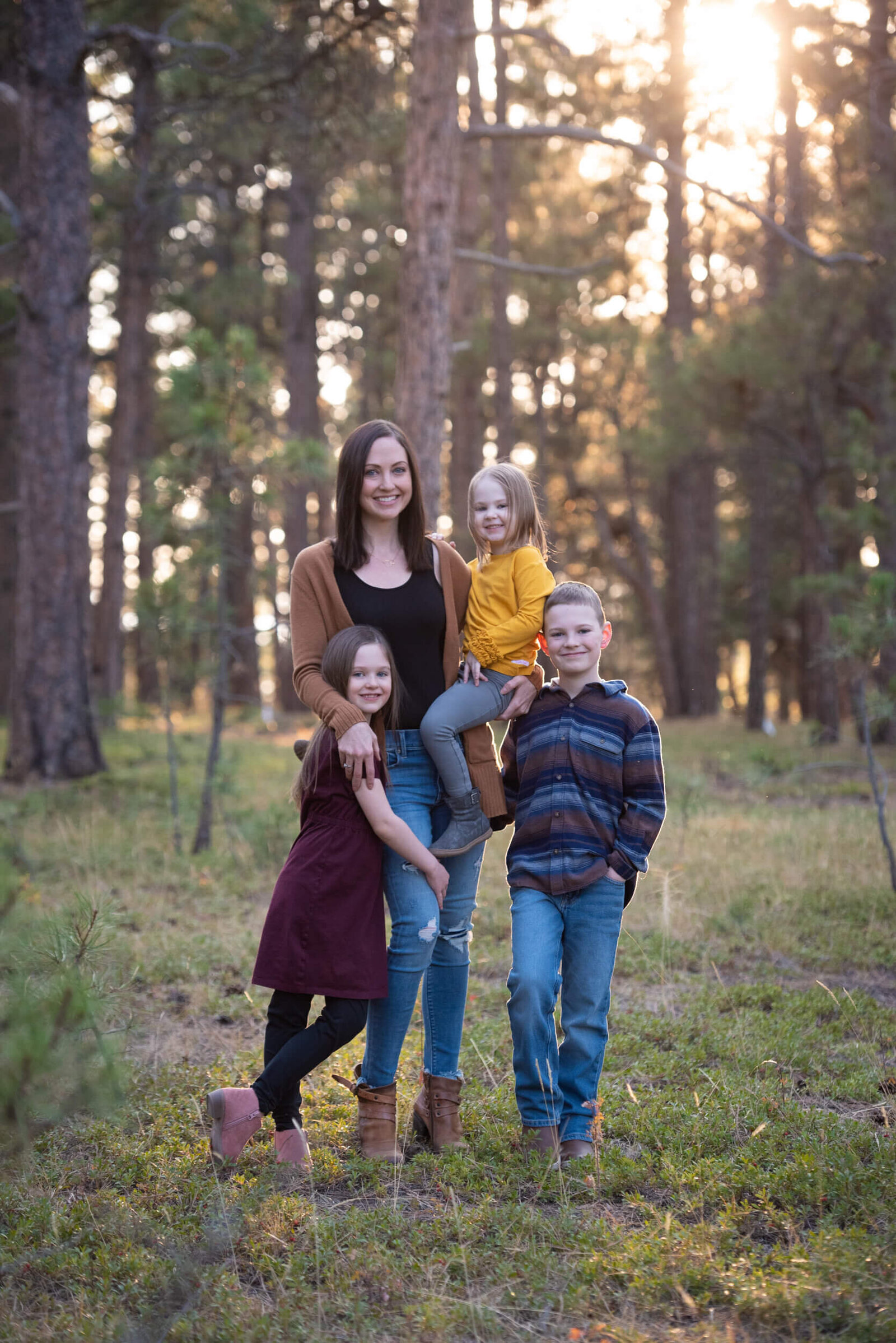 A mother in a black shirt stands in a forest with her three toddler children smiling for a Denver family photographer