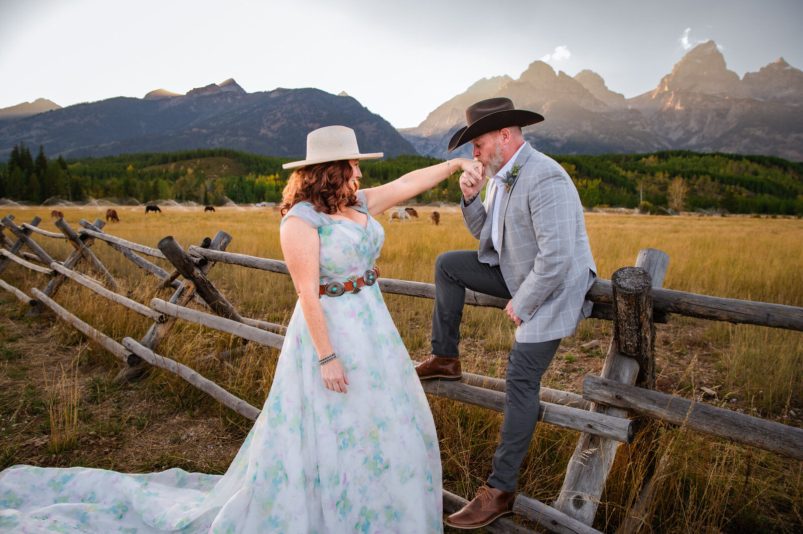 Groom sits on fence and kisses brides hand in Grand teton national park. Both are wearing cowboy hats and bride is in a blue and white dress.
