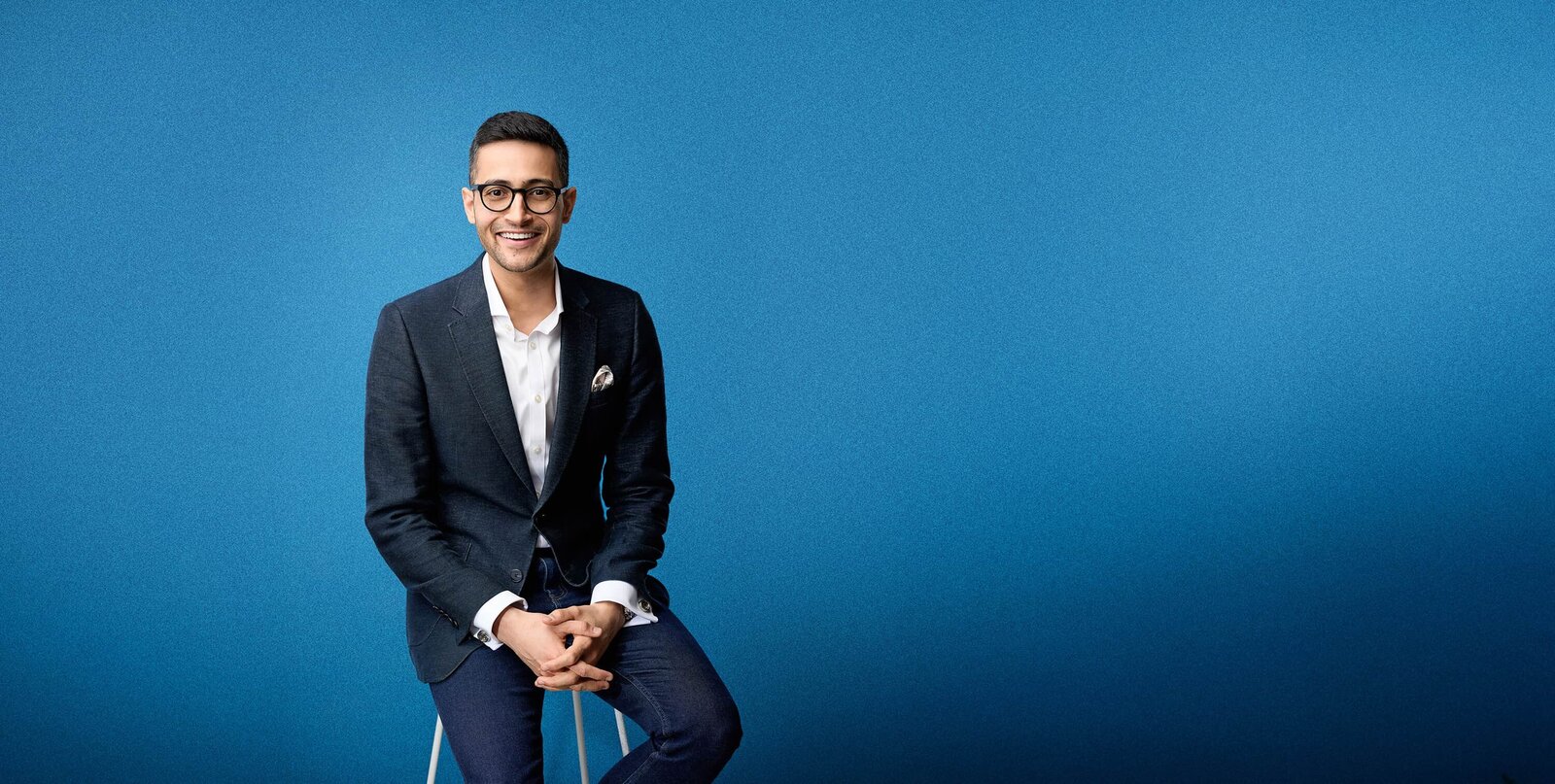 Professional photo of Yash with a blue gritty background. He's on a stool and is smiling to camera wearing jeans, white shirt and blue jacket.