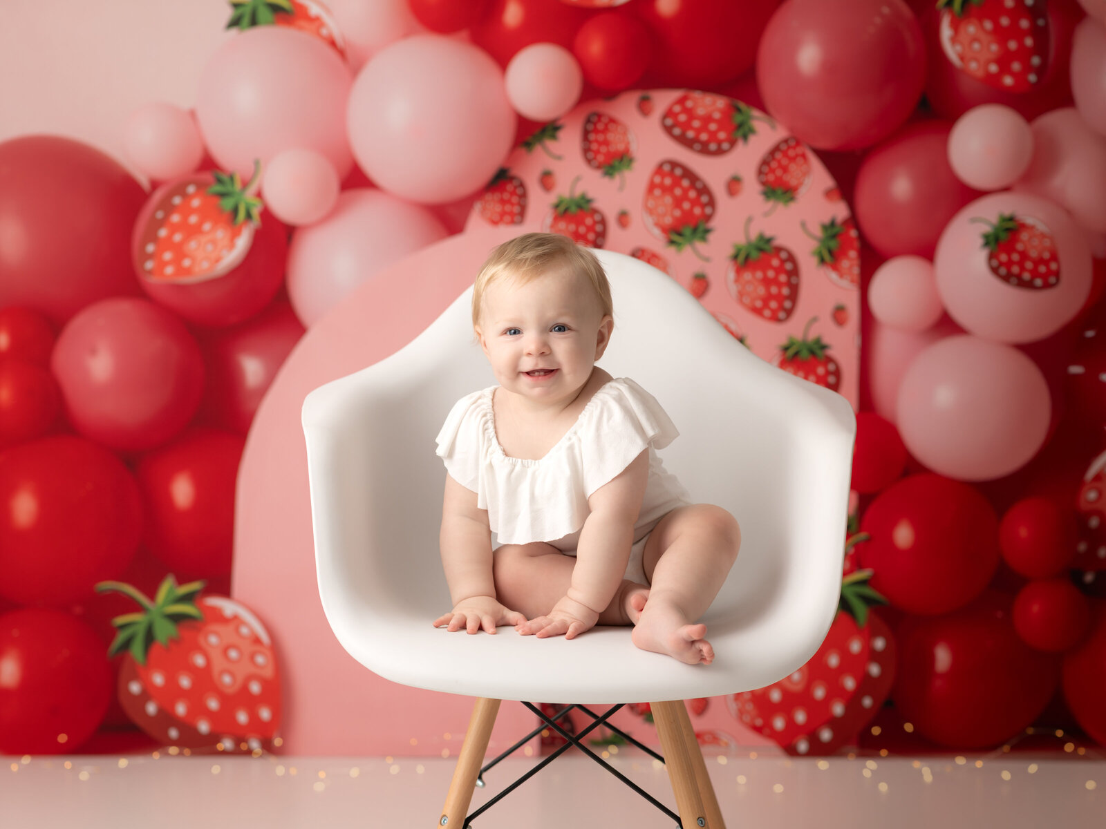 one year old girl on white onesie sitting in white chair with strawberry theme backdrop