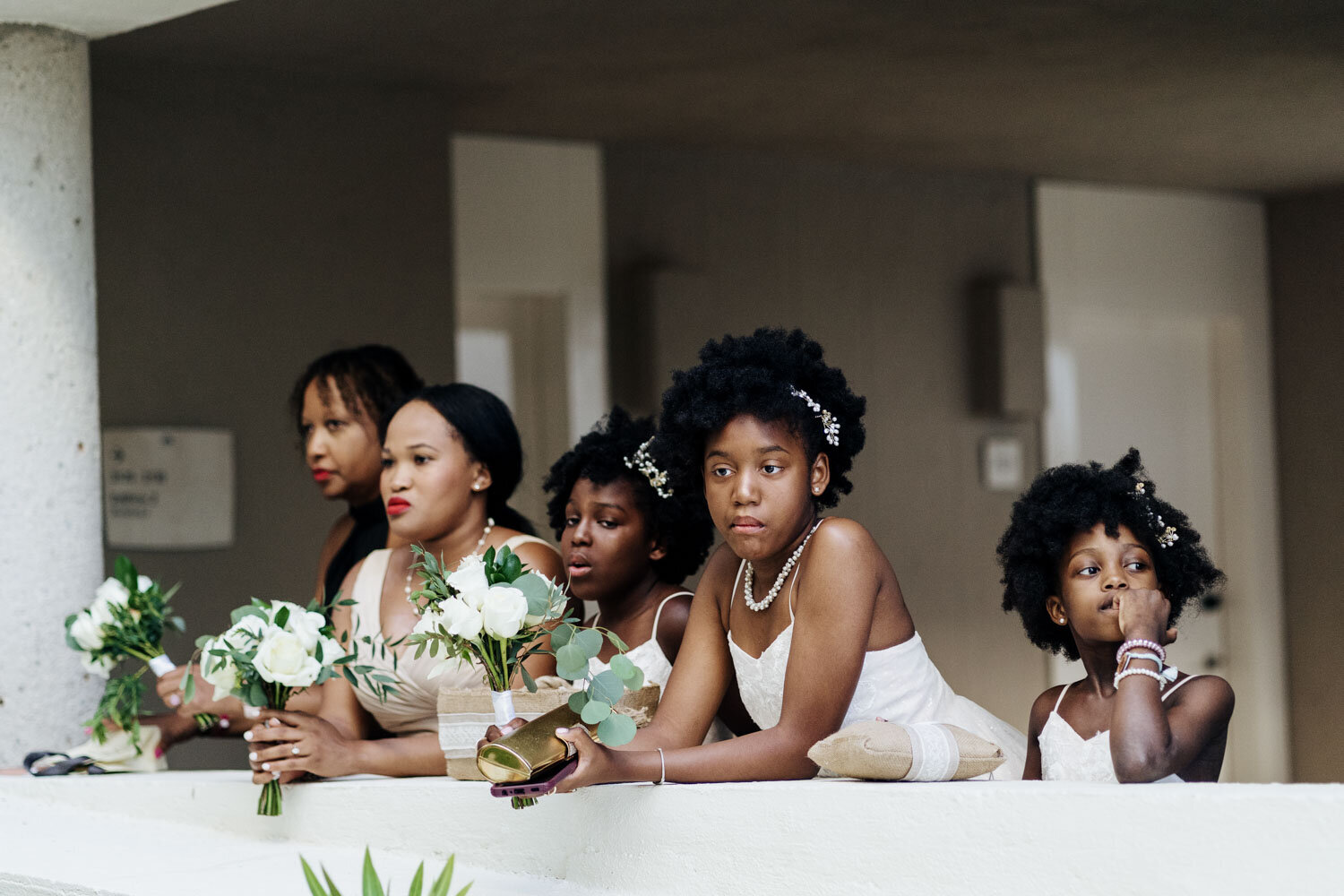 Bridesmaids in white, holding bouquets, watching from a balcony