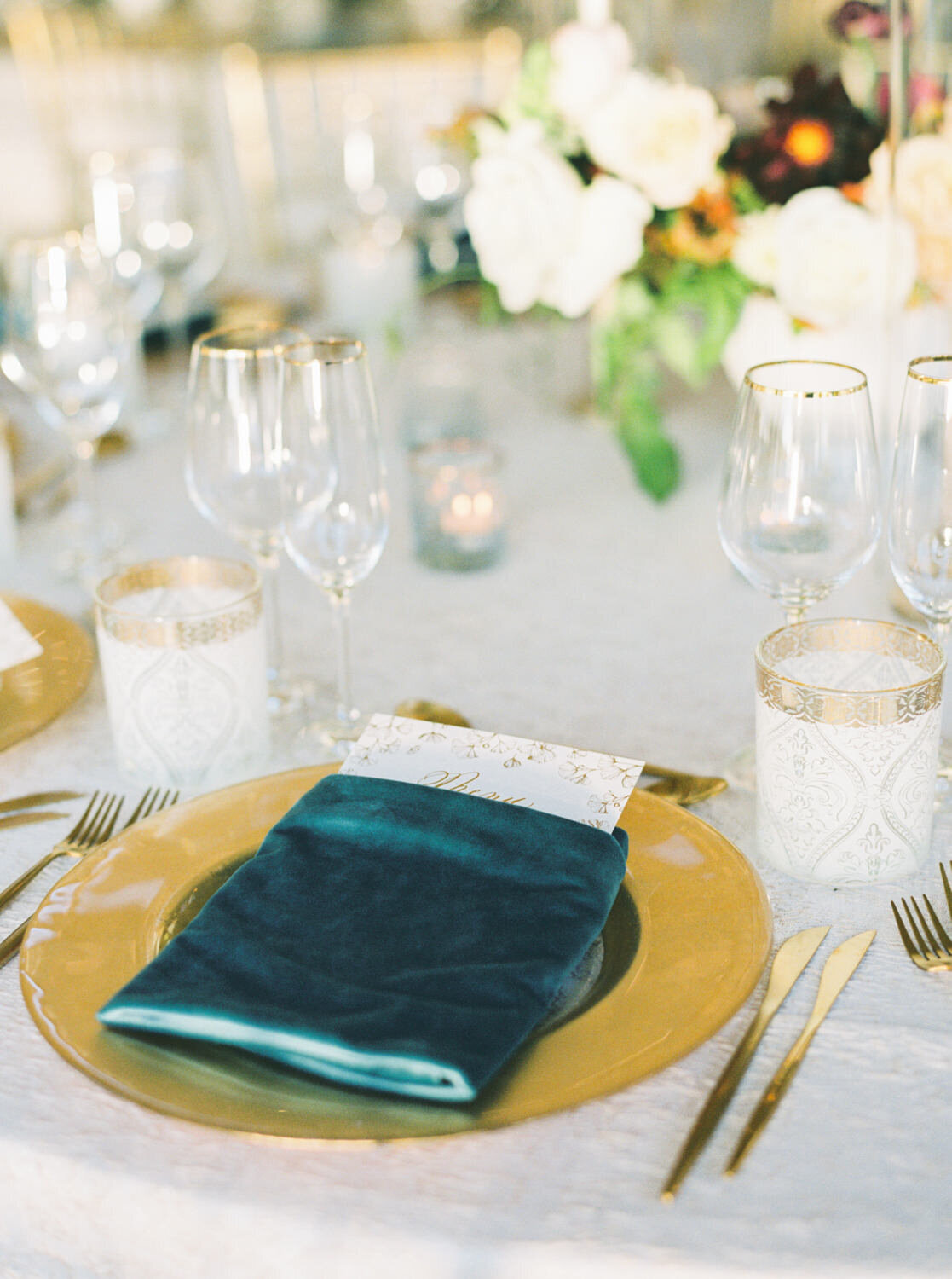 A blue velvet napkin holds a menu on a gold charger. Luxury glassware and candles in the background.