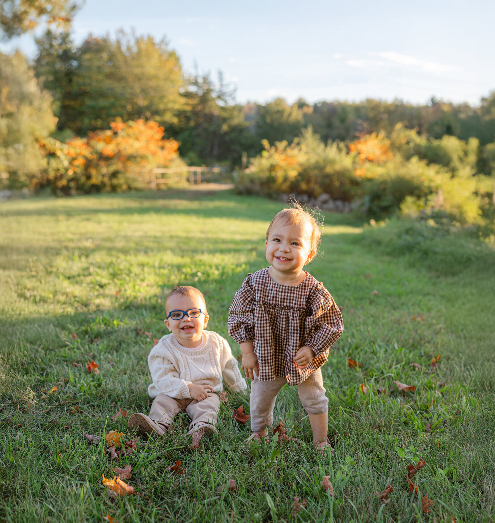 Twin toddlers smile in a grassy field surrounded by early fall colors, photographed by a Worcester family photographer.