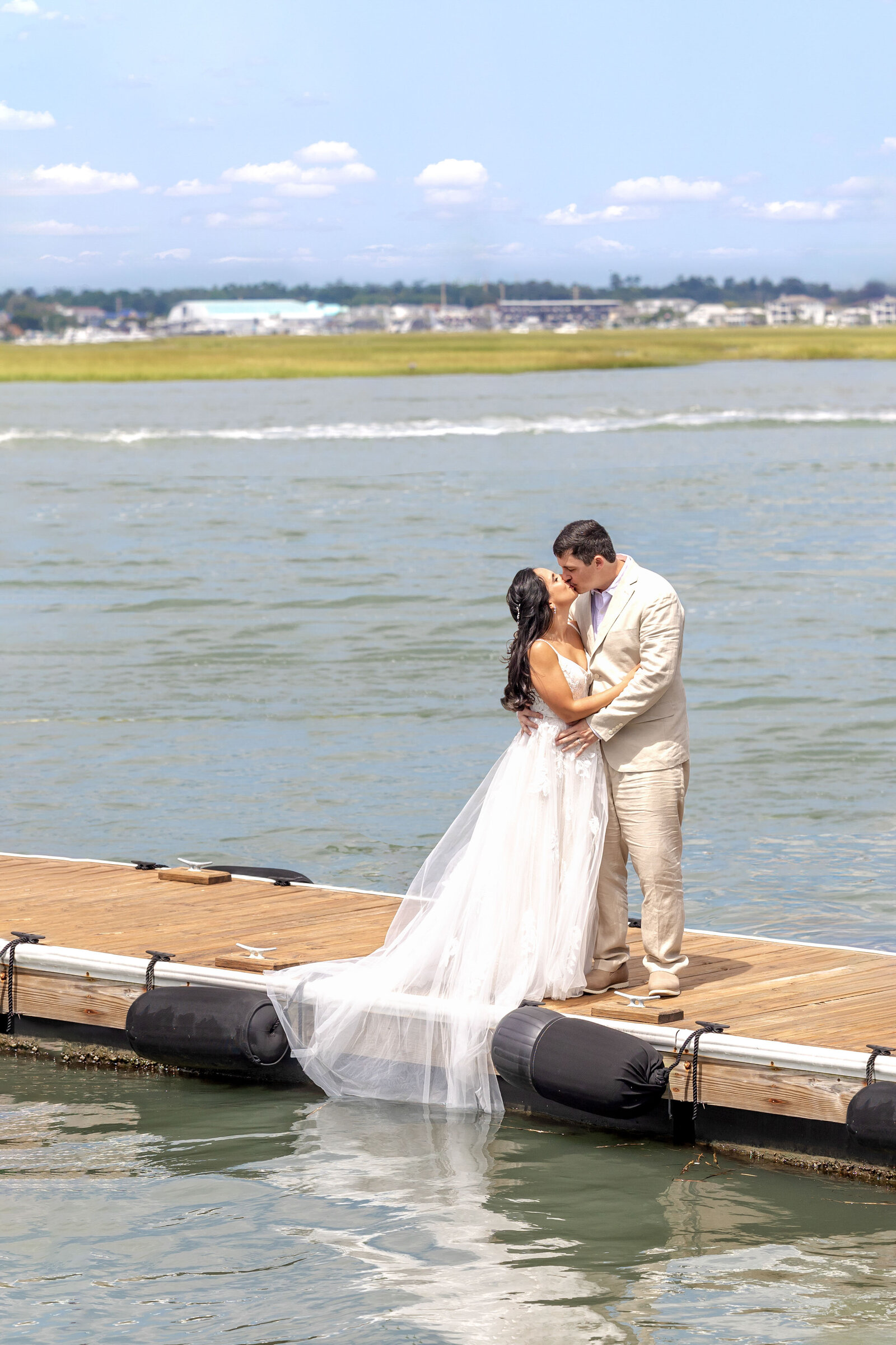 The bride and groom share a romantic kiss on a dock overlooking the water at a Wrightsville Beach, NC wedding, blending coastal charm with authentic and intimate moments.
