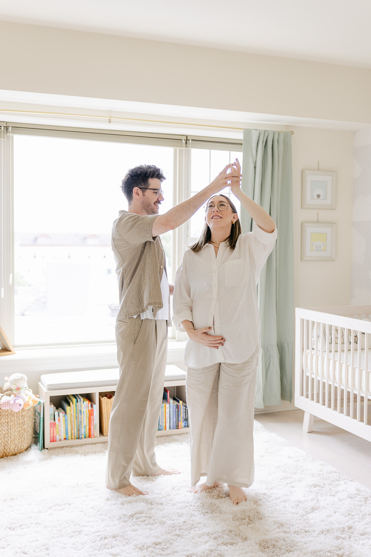 A couple dances playfully in their sunlit nursery, the expectant mother smiling as she’s twirled by her partner. Surrounded by soft neutral tones, a wooden crib, and a cozy book nook, this moment captures the anticipation, connection, and quiet joy of preparing for baby. A candid, love-filled highlight from a maternity photo session in Greenwich, CT.