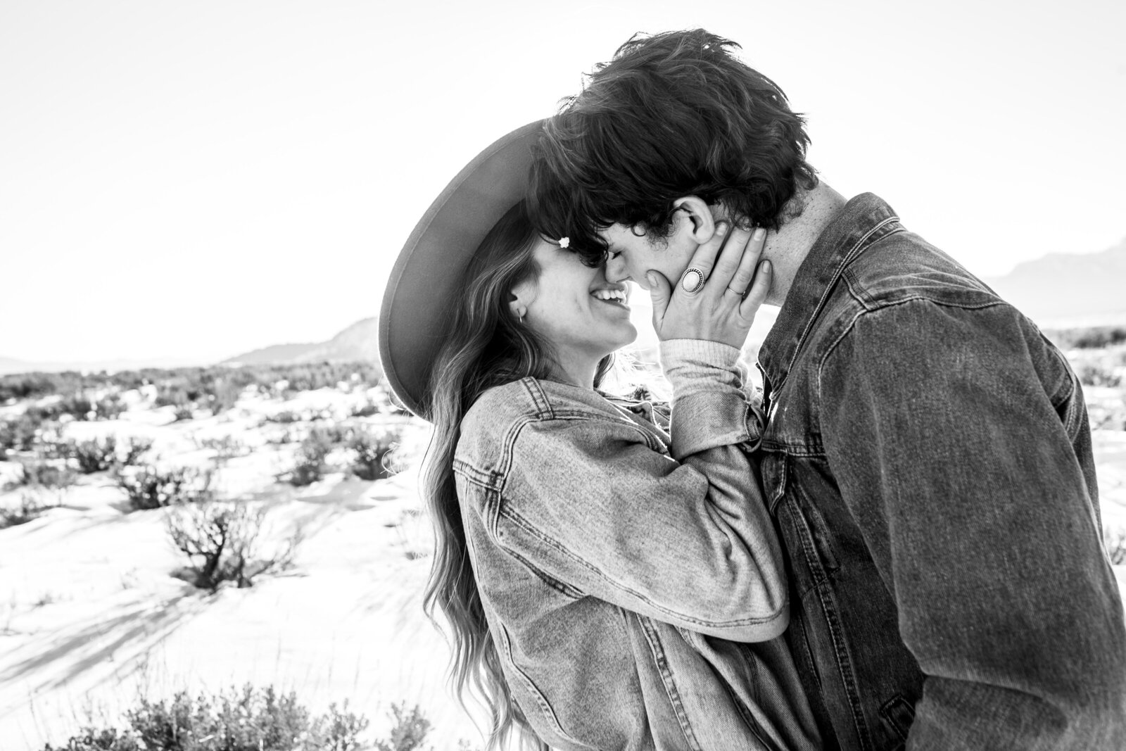 black and white image of engaged couple about to kiss with the man leaning into the woman as she holds his face and smile, they are both wearing denim and she is wearing a wide brimmed hat