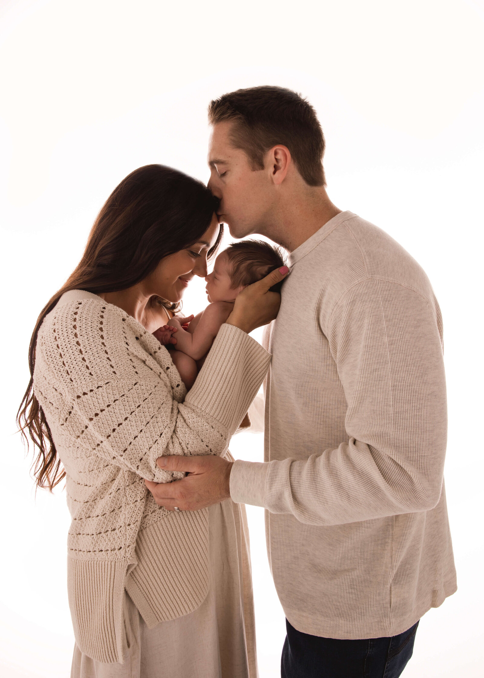 Couple embraced while holding baby. Husband kissing wifes forehead in studio by Ashley Nicole.