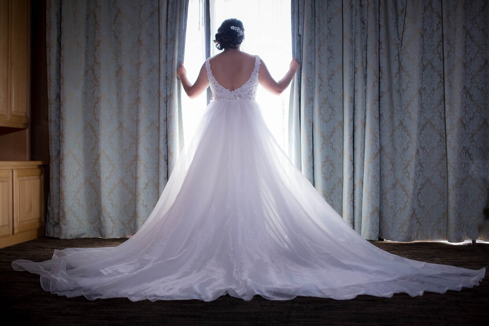Bride pushes away curtains to look outside with large train of dress spread out behind her.  Photo taken by Philippe Studio Pro, Sacramento wedding photography studio.
