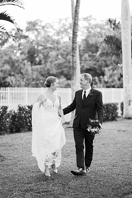 Black and white image of bride and groom walking across lawn, groom is holding brides bouquet for her while bride holds her dress up so she can walk.