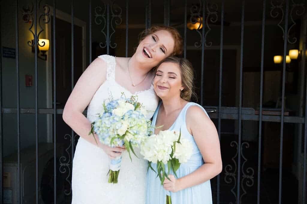 A bride and a bridesmaid wearing a light blue colored dress smile and stand close together, each holding a bouquet of white and blue flowers, in front of a decorative metal gate in Ruston, Louisiana.