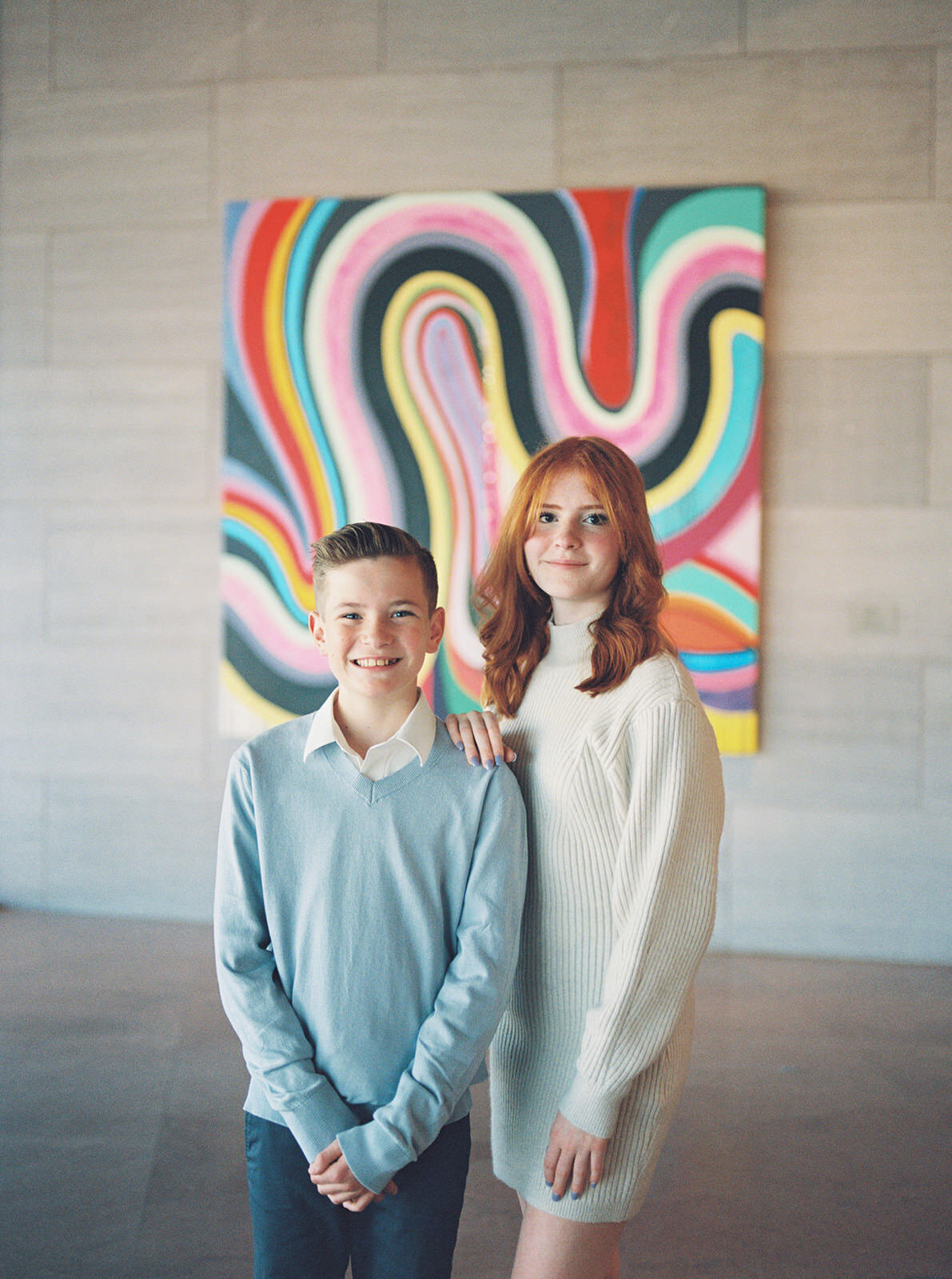 "A vibrant portrait of a red-haired teenage girl and her younger brother, posed in front of a colorful abstract painting in a modern gallery."