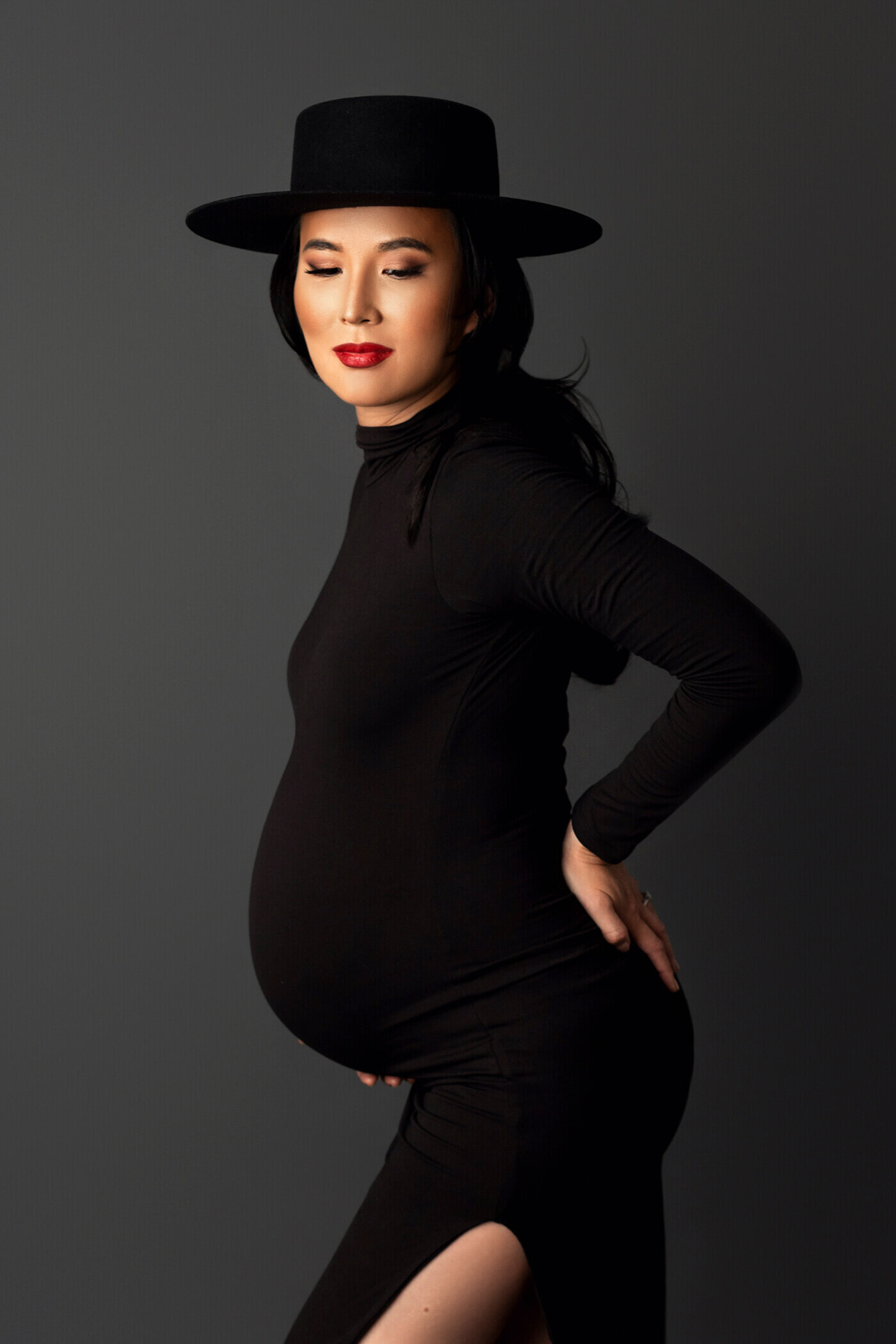 A stylish pregnant woman poses confidently against a dark background, wearing a form-fitting black dress with a high neckline and long sleeves. The dress features a thigh-high slit, revealing her leg. She accessorizes with a bold black wide-brimmed hat and wears bright red lipstick, adding a touch of sophistication. The woman's eyes are closed, and her hand rests on her hip, emphasizing her baby bump. The image captures her elegance and poise during pregnancy.