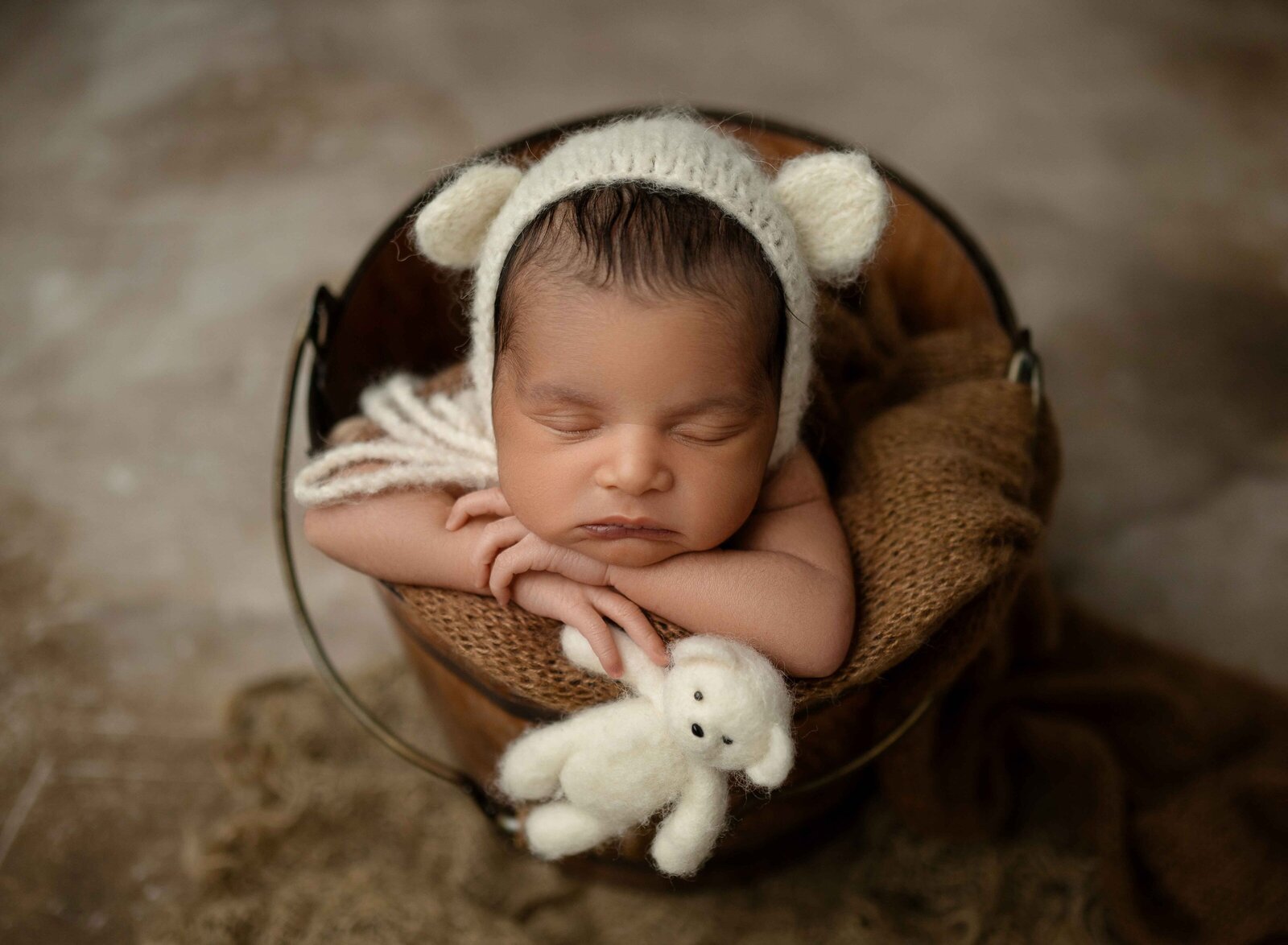 Baby boy wearing a bear bonnet and sleeping in a brown bucket with a felted teddy bear.