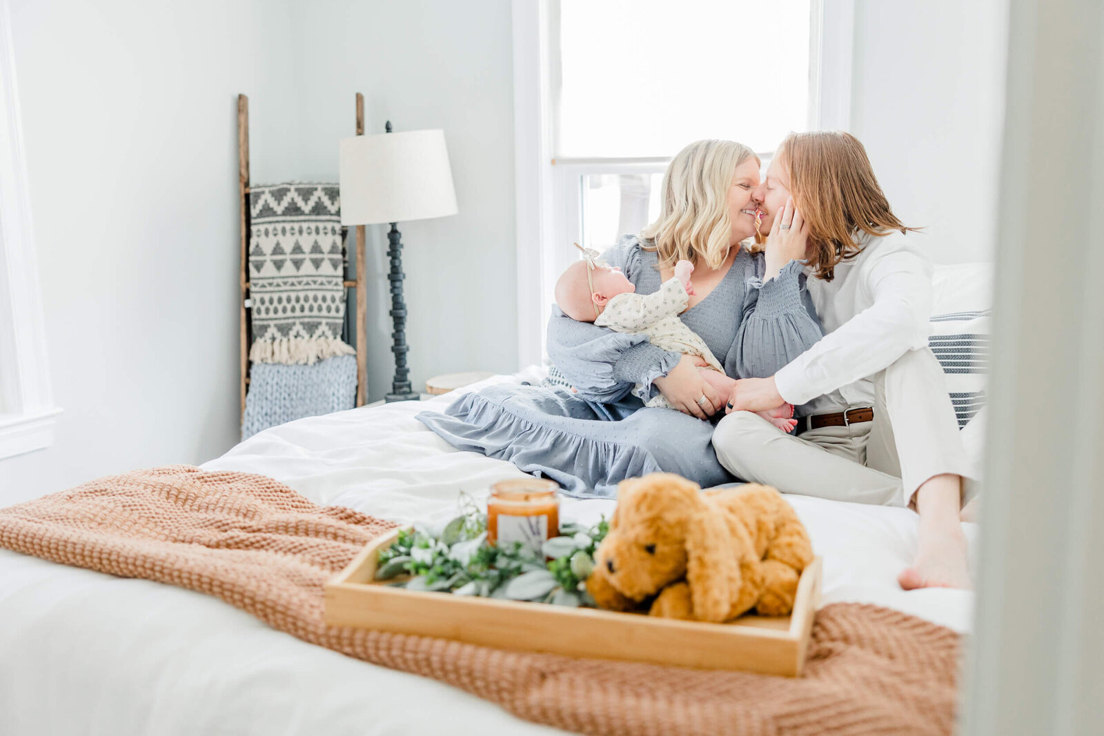 Mom and dad lean in for a kiss while mom holds the newborn baby on the bed