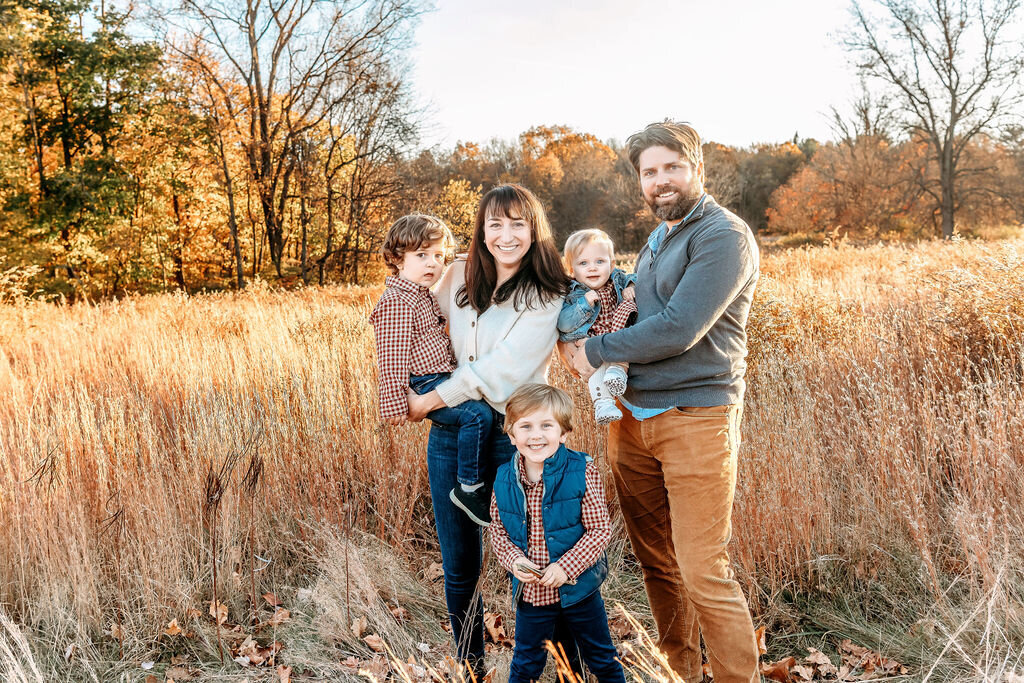 Connecticut Family Photographer documents a family of five posing in a golden autumn field, with parents and three children wearing coordinated fall outfits among tall grasses and colorful foliage