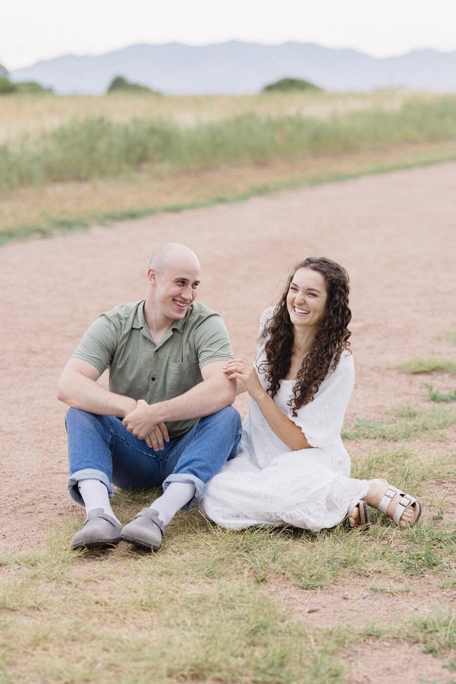 A couple sit on a red dirt path laughing with one another.