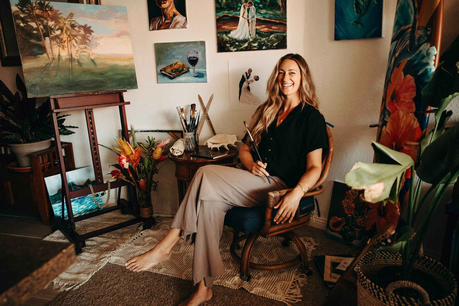 Artist Jules Trowbridge smiles while seated, surrounded by her art and supplies in the studio