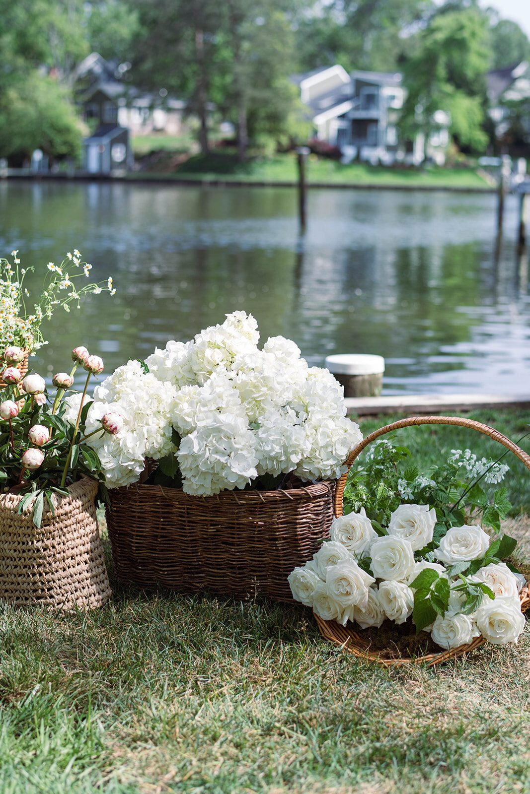 Wicker baskets filled with white garden roses, white hydrangeas, daisies, and blush peonies with scenic waterfront in the background.