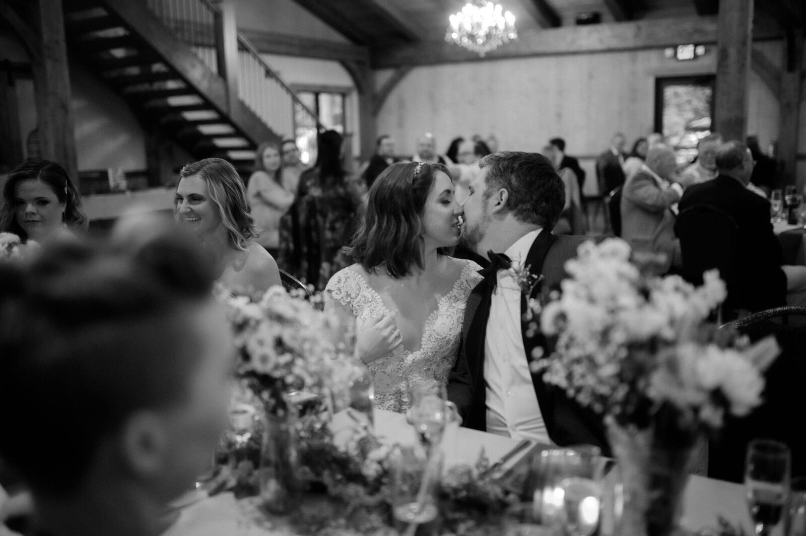 bride and groom sitting at table kissing at wedding reception, guests clapping