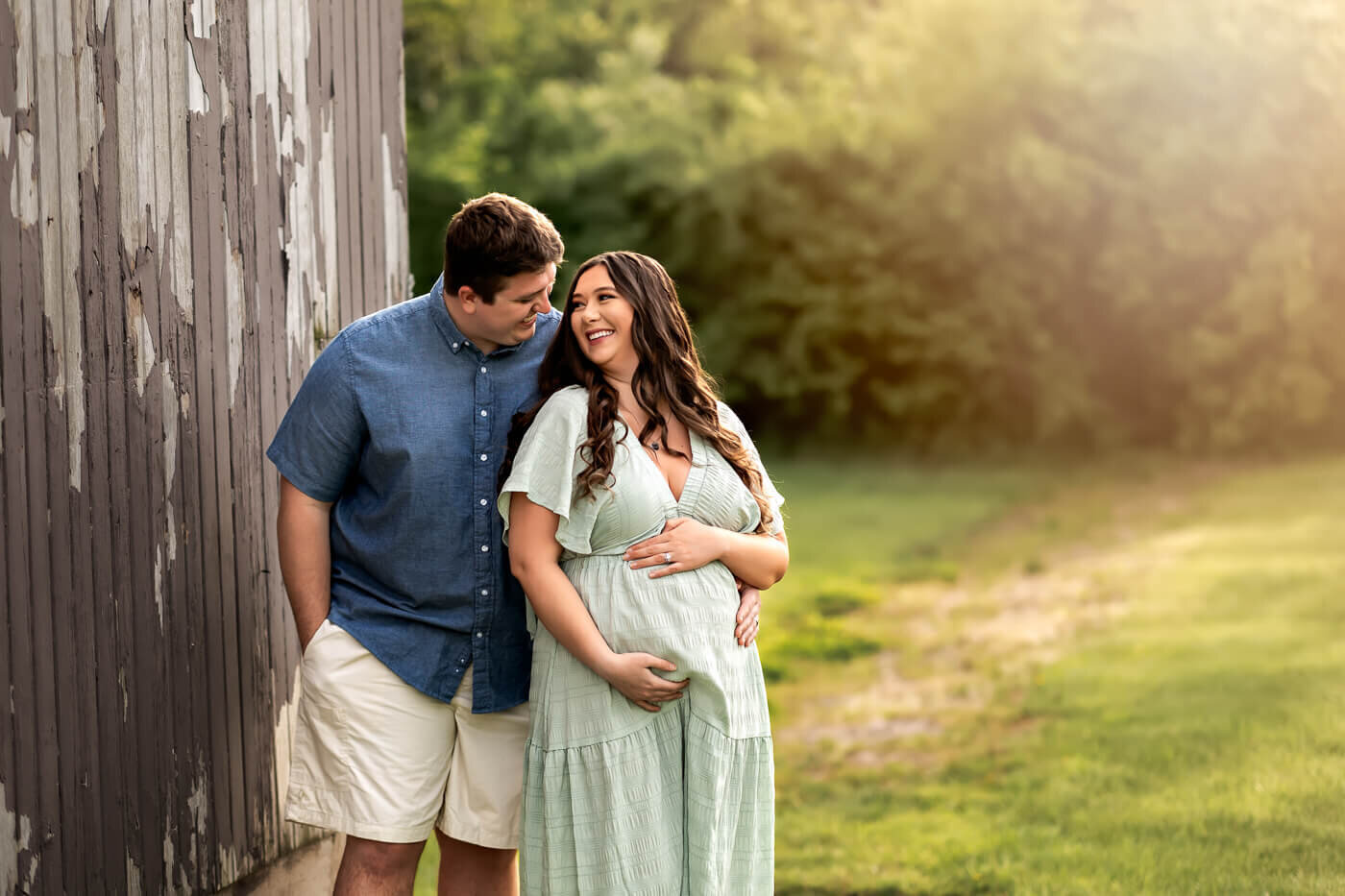 A smiling couple stands by a weathered wooden wall outdoors; the woman, expecting, cradles her belly as her partner leans close. Surrounded by green grass and trees in soft sunlight, this tender moment is perfect for maternity portraits.