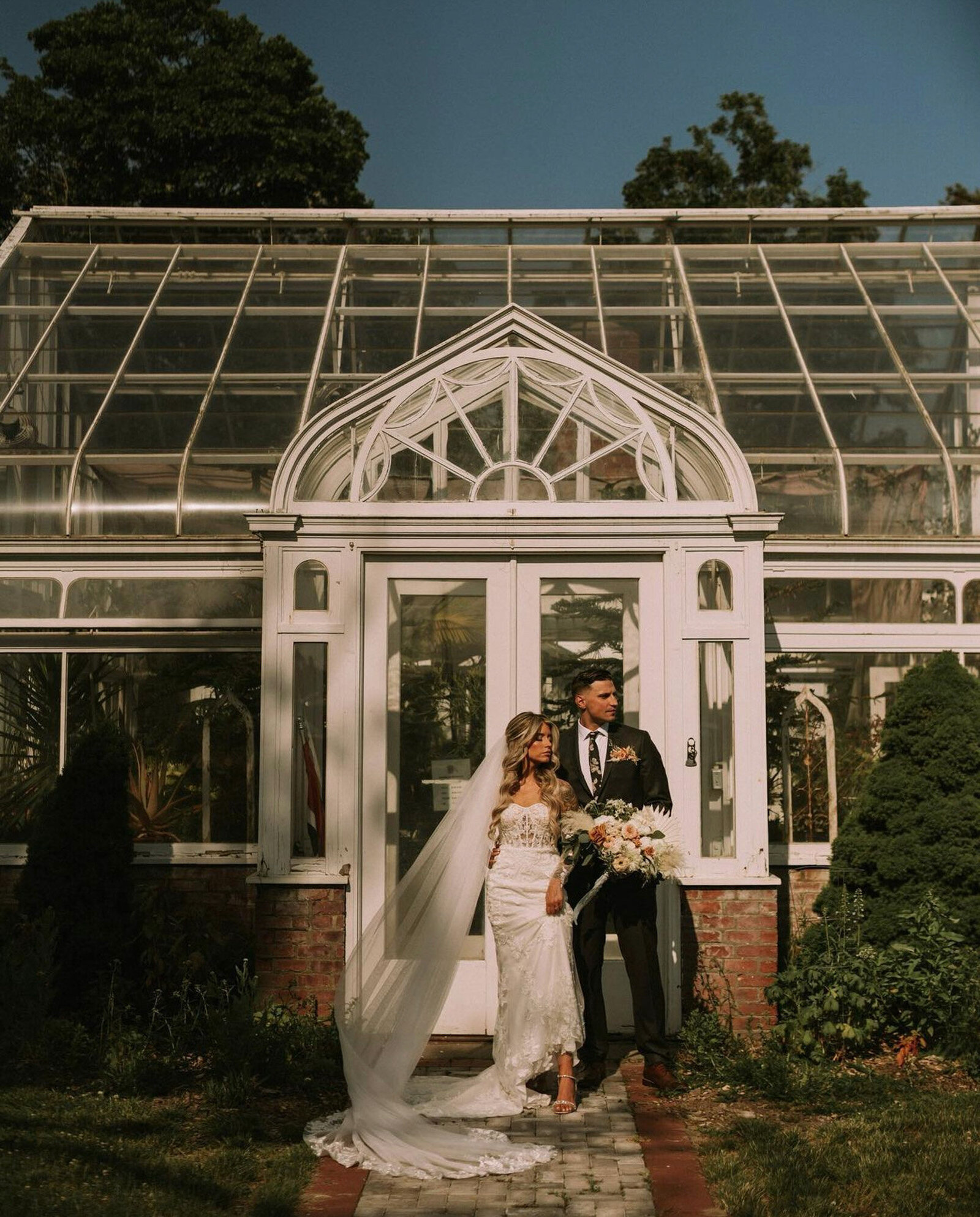 bride and groom standing arm-in-arm on a brick path surrounded by greenery in front of a glass greenhouse