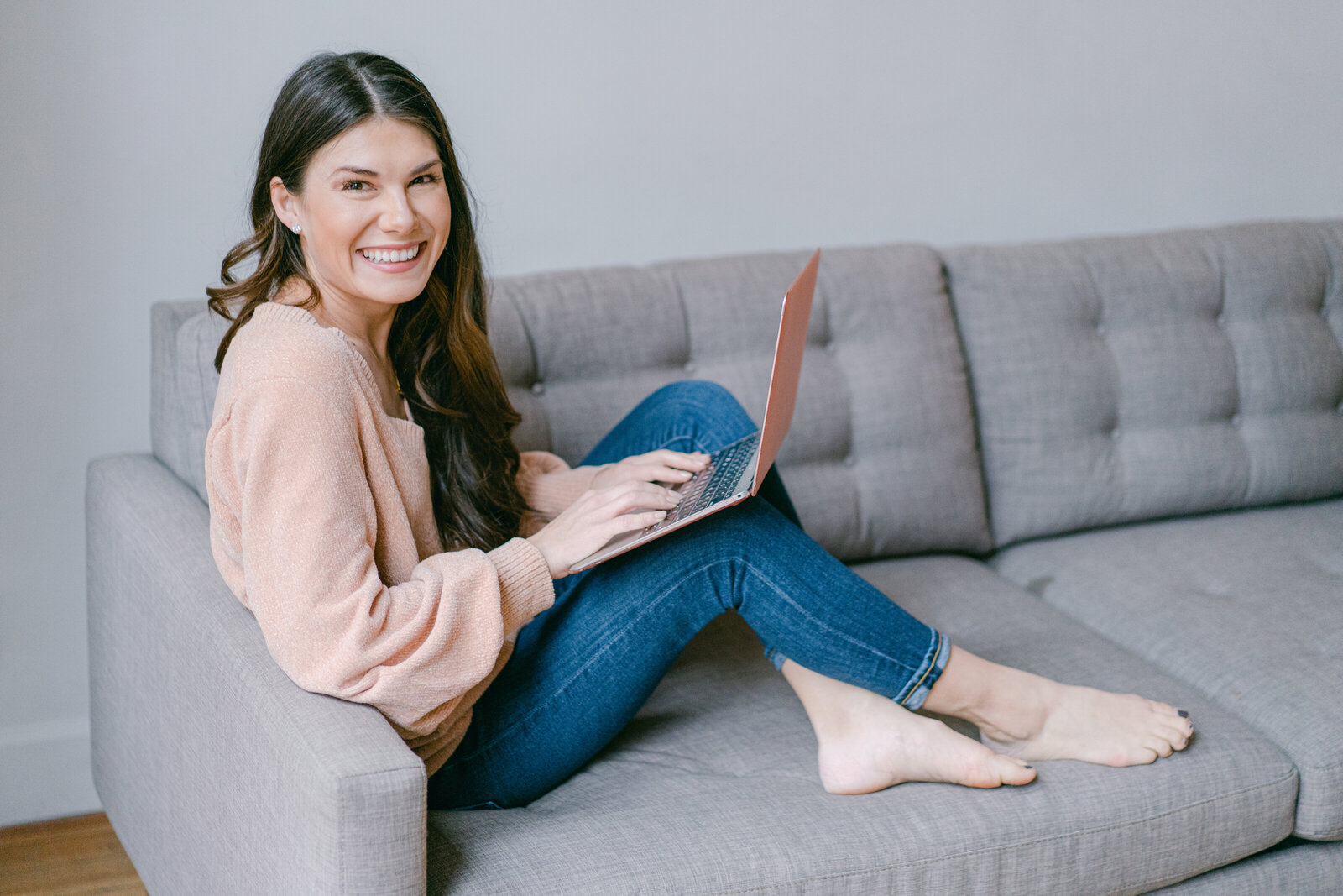 Woman sits on grey couch with laptop smiling at camera