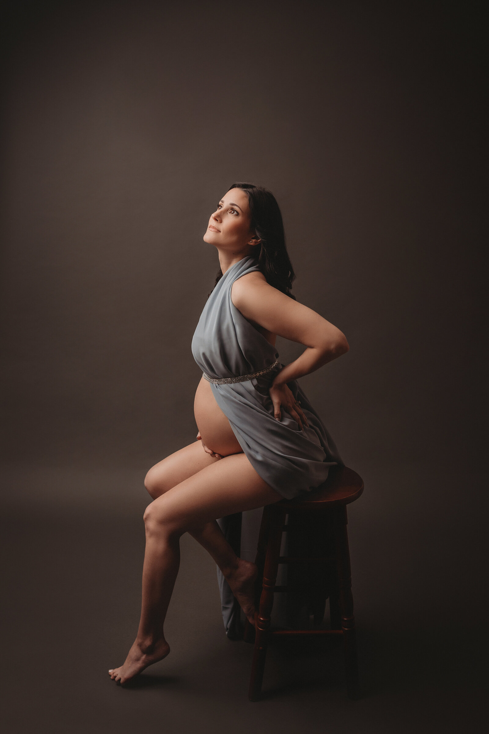 35 week pregnant mother to be at Marietta GA maternity studio sitting on wooden stool dressed in blue chiffon holding baby bump and leaning up towards and looking at light on dark gray backdrop