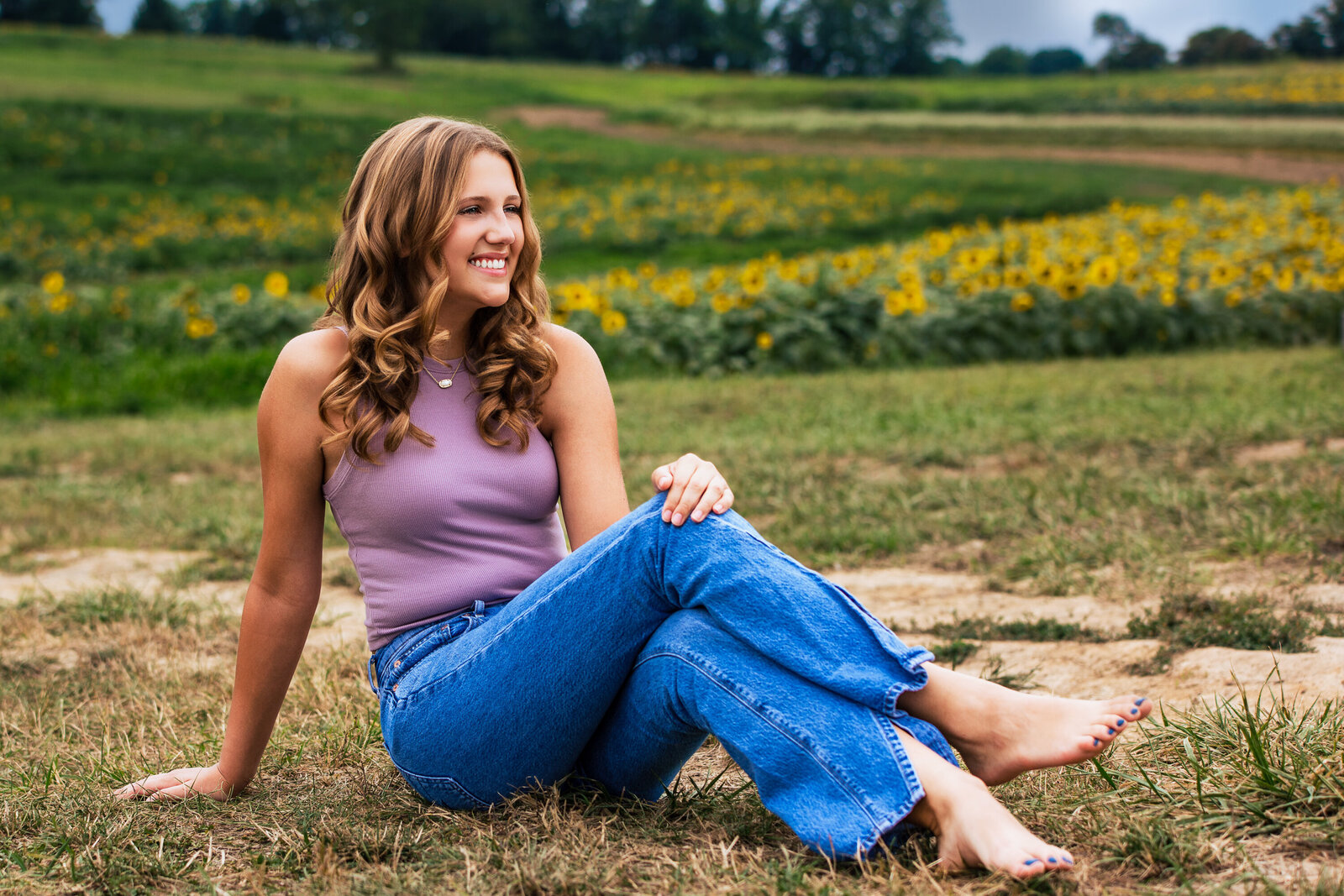 A senior girl poses with her legs crossed looking off into the distance in a beautiful Golden sunflower field.
