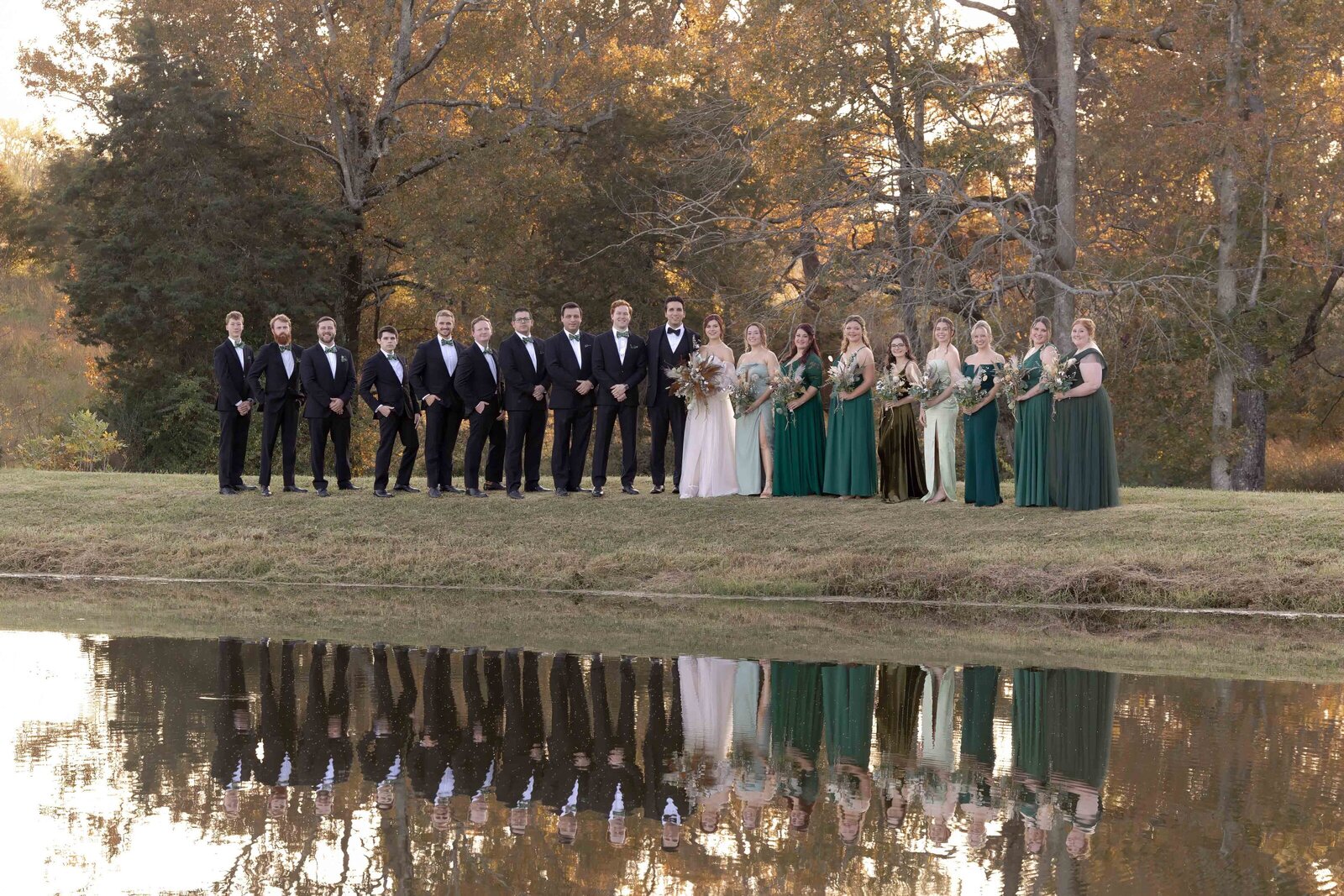 A large wedding party poses by a reflective pond during an autumn wedding in Boone, NC, surrounded by vibrant fall foliage and rustic elegance, creating a timeless and picturesque scene.