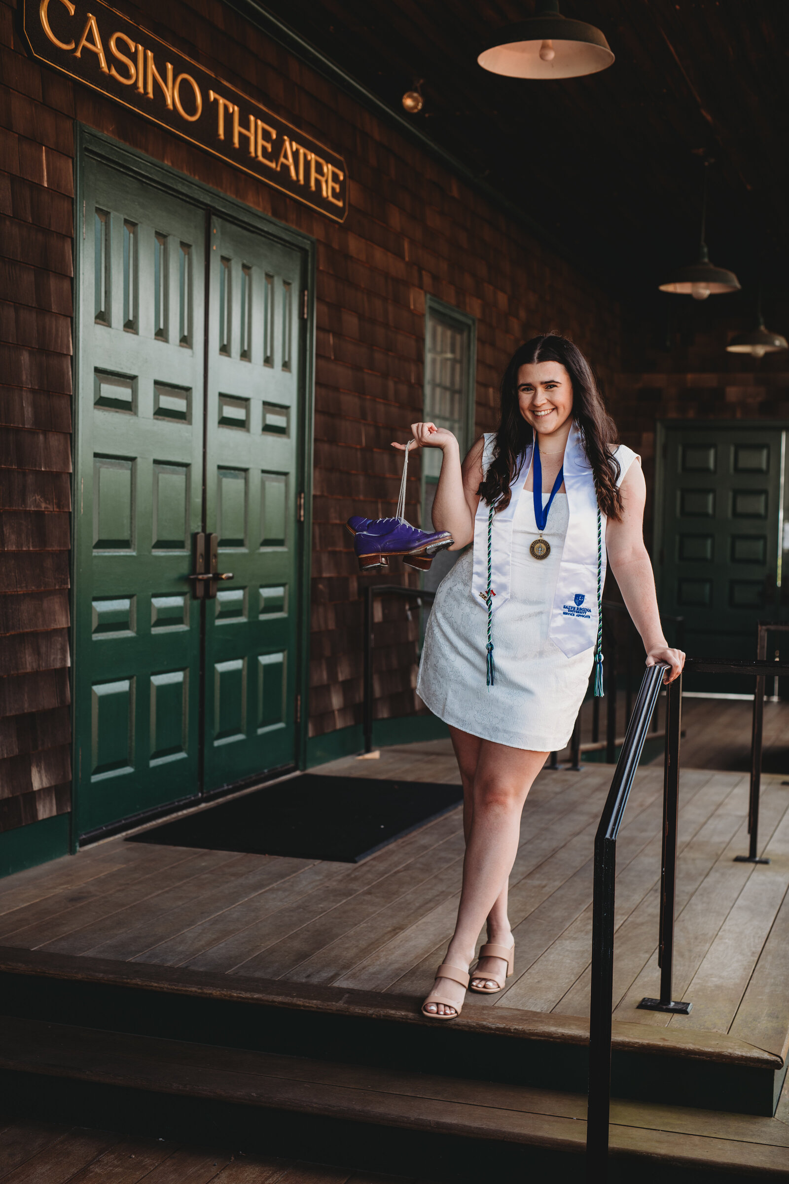 A proud graduate of Salve Regina University poses on the steps of the historic Casino Theatre, holding a symbolic purple shoe and wearing academic honors. Captured in Newport, Rhode Island, by Nicole Mele Photography, specializing in timeless college graduation portraits.